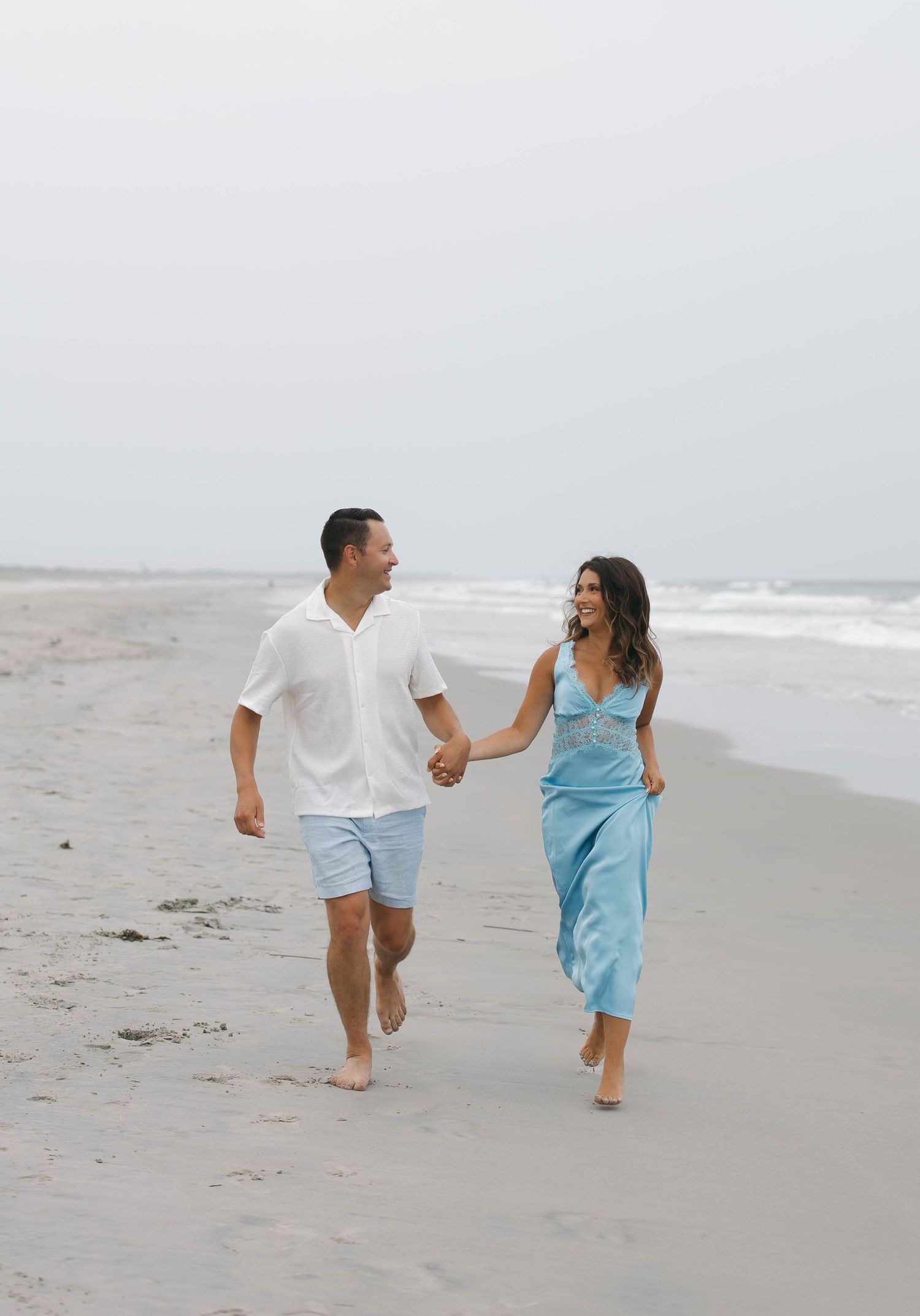 A playful couple runs along the beach wearing coordinated blue and white outfits.