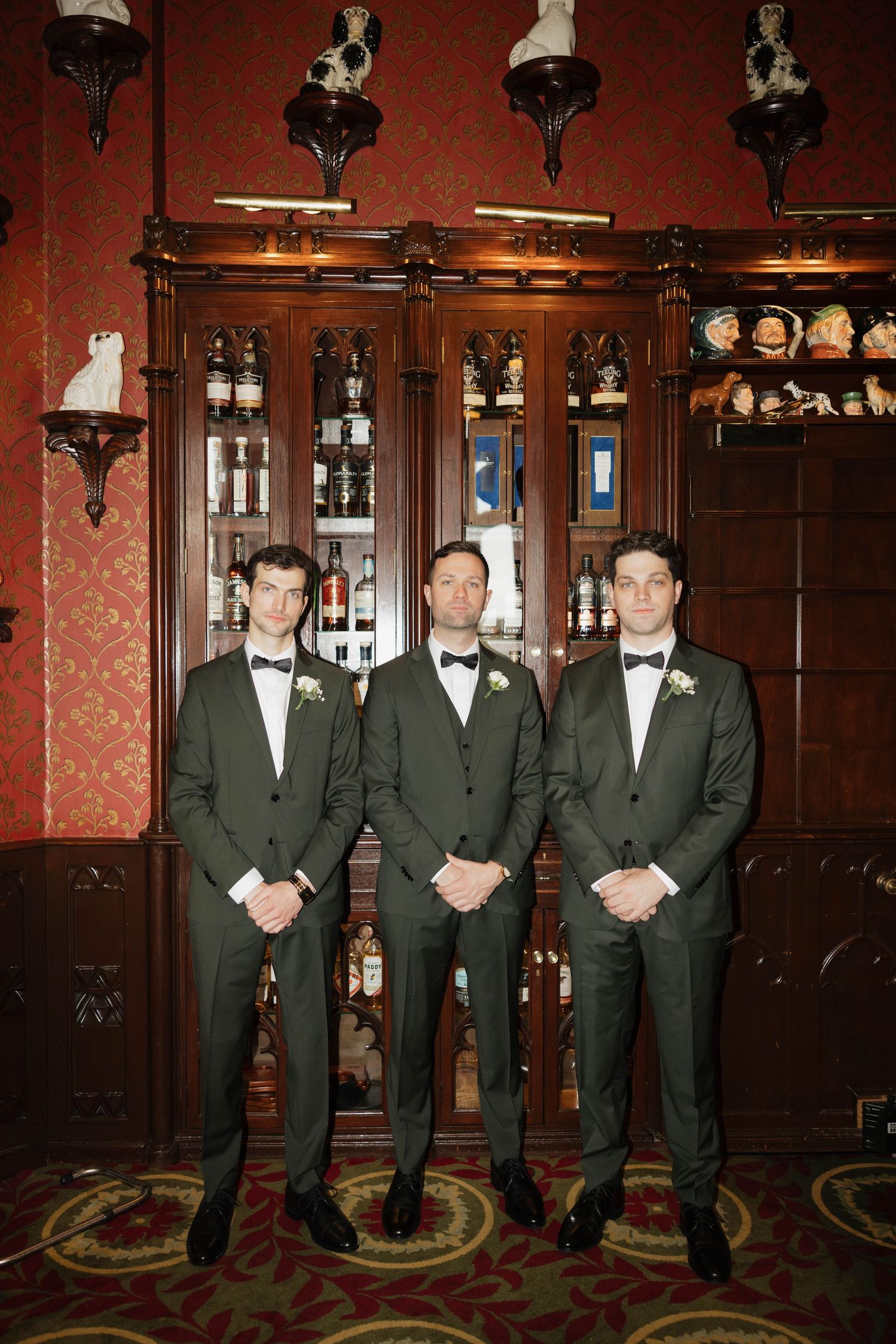 Wedding party members stand in formation wearing black tuxedos in an elegant bar setting.