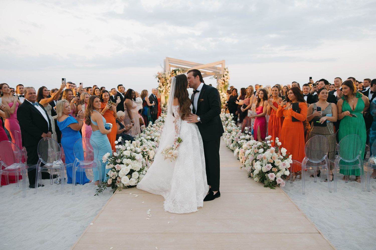 Wedding guests gather on a sandy beach during a seaside ceremony with elegant decor and floral arrangements.