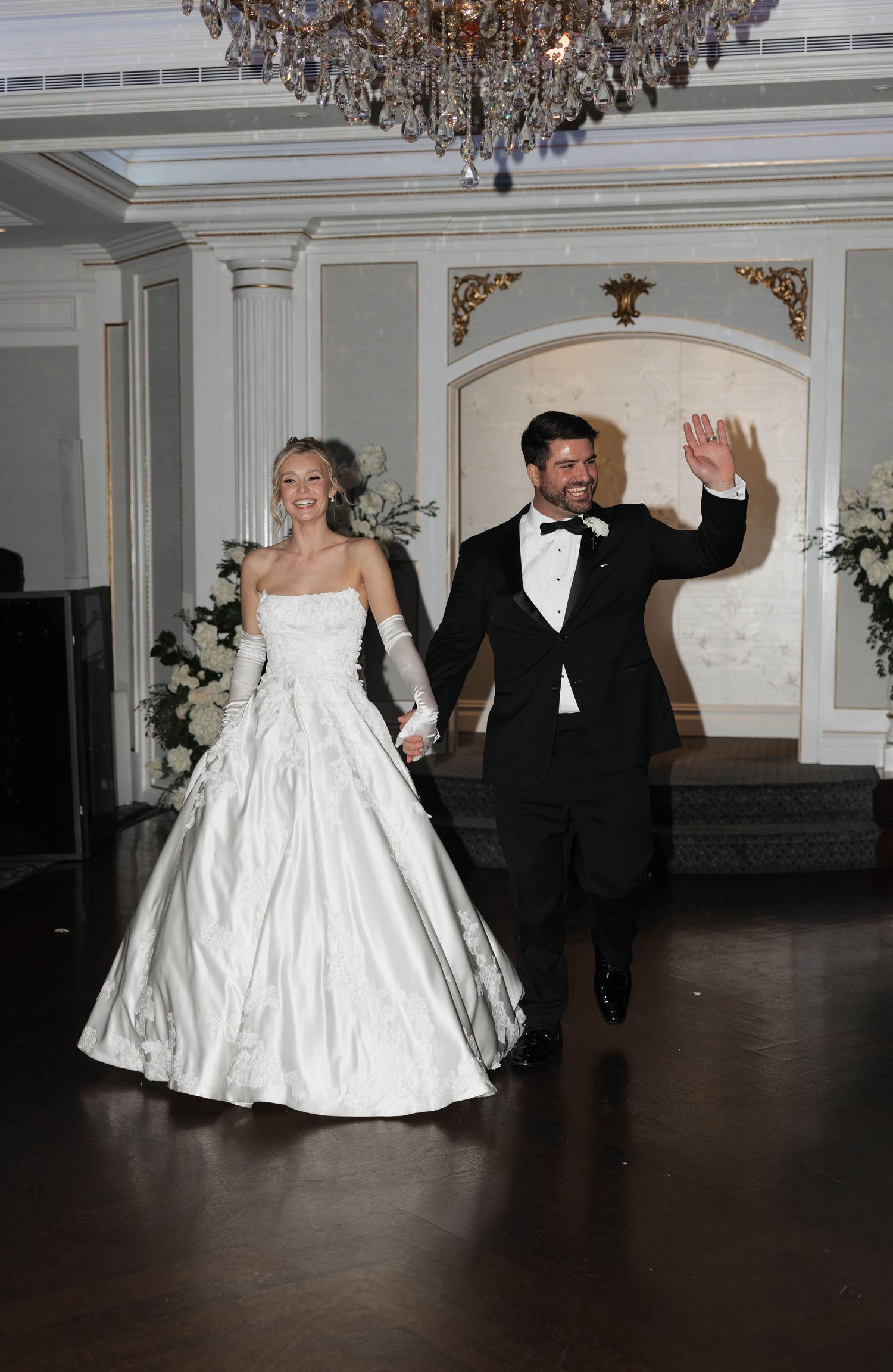 Wedding couple making joyful entrance into reception hall under ornate chandelier.