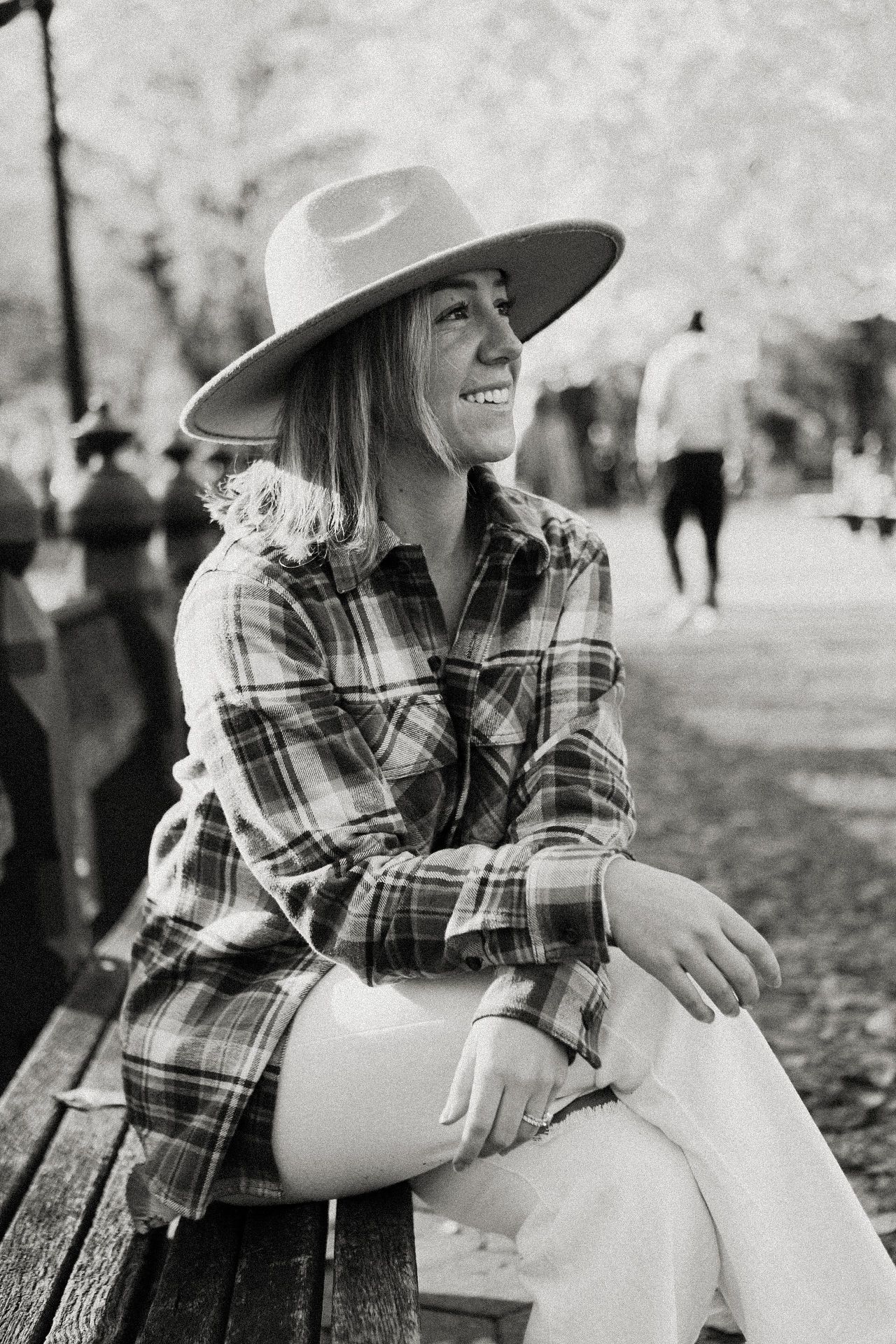 Black and white portrait of someone in plaid and a wide-brim hat sitting outdoors.