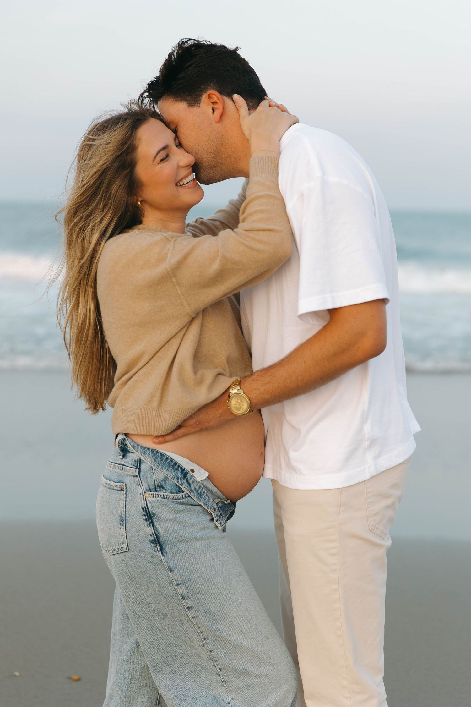 A tender moment between a couple on the beach, dressed casually in light colors against an ocean backdrop.