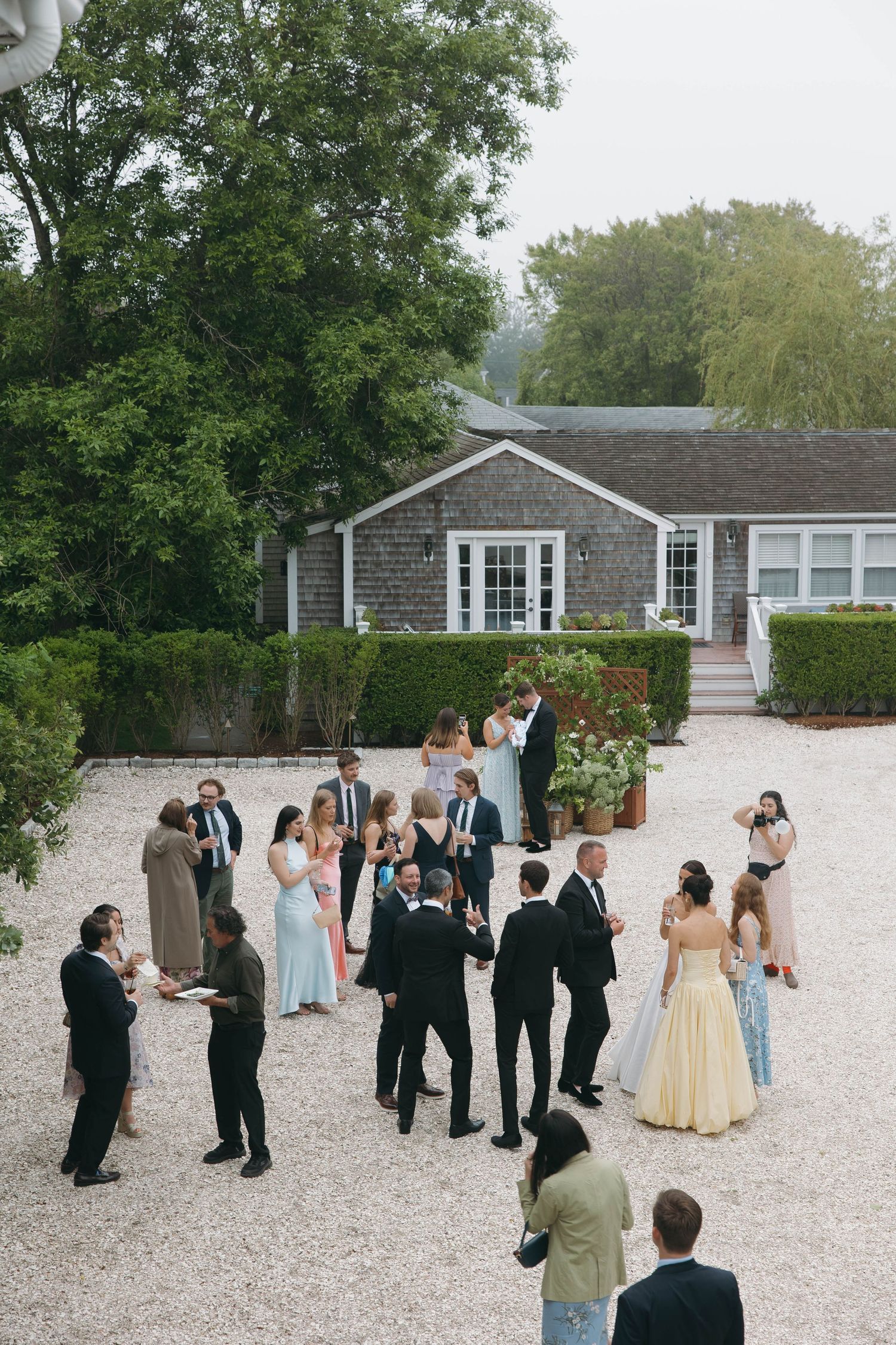 Wedding guests mingling on a white gravel courtyard outside a gray shingled building.