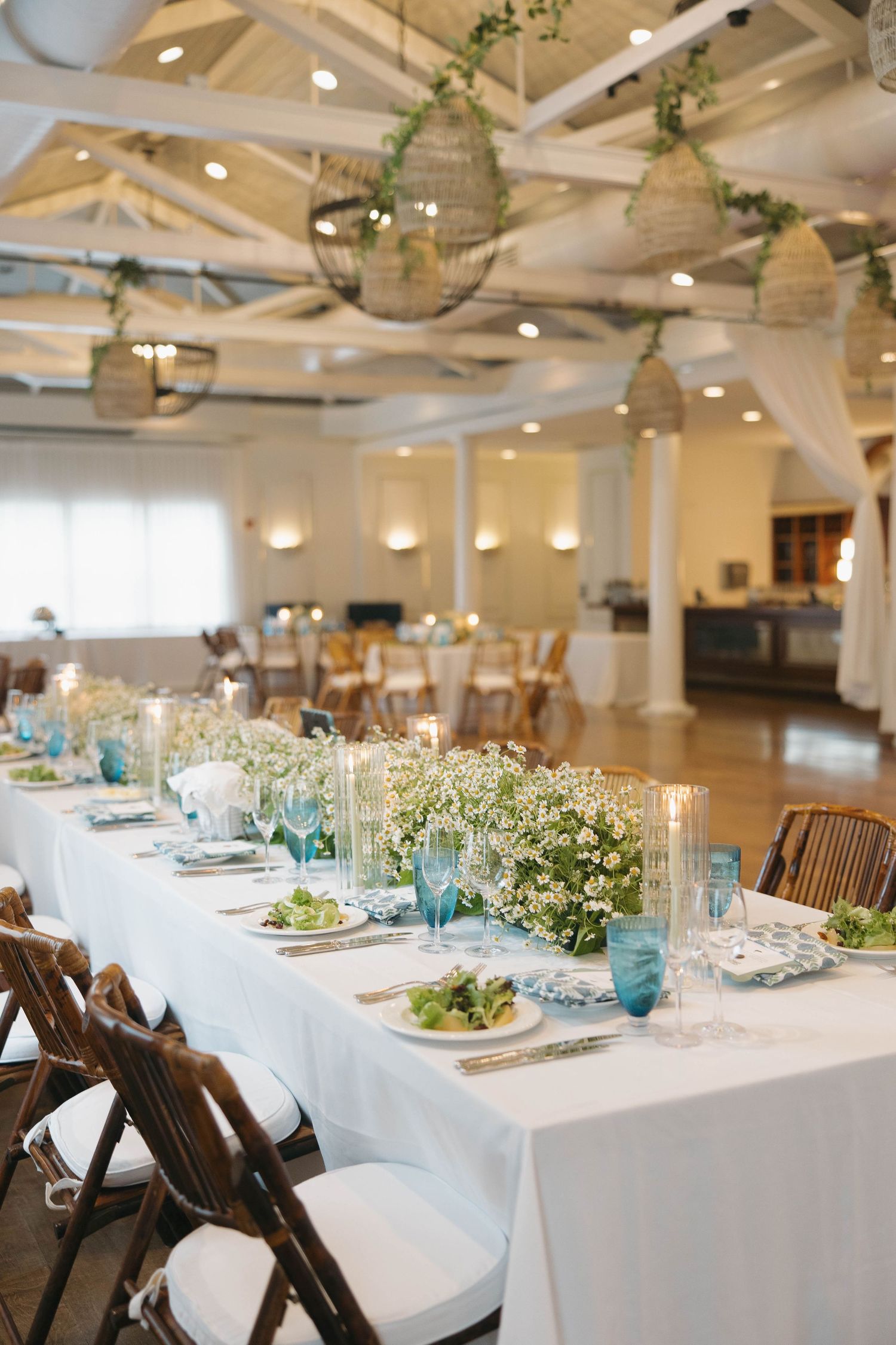 Elegant wedding reception venue with white tables, wooden chairs, and hanging lanterns in a bright white room.