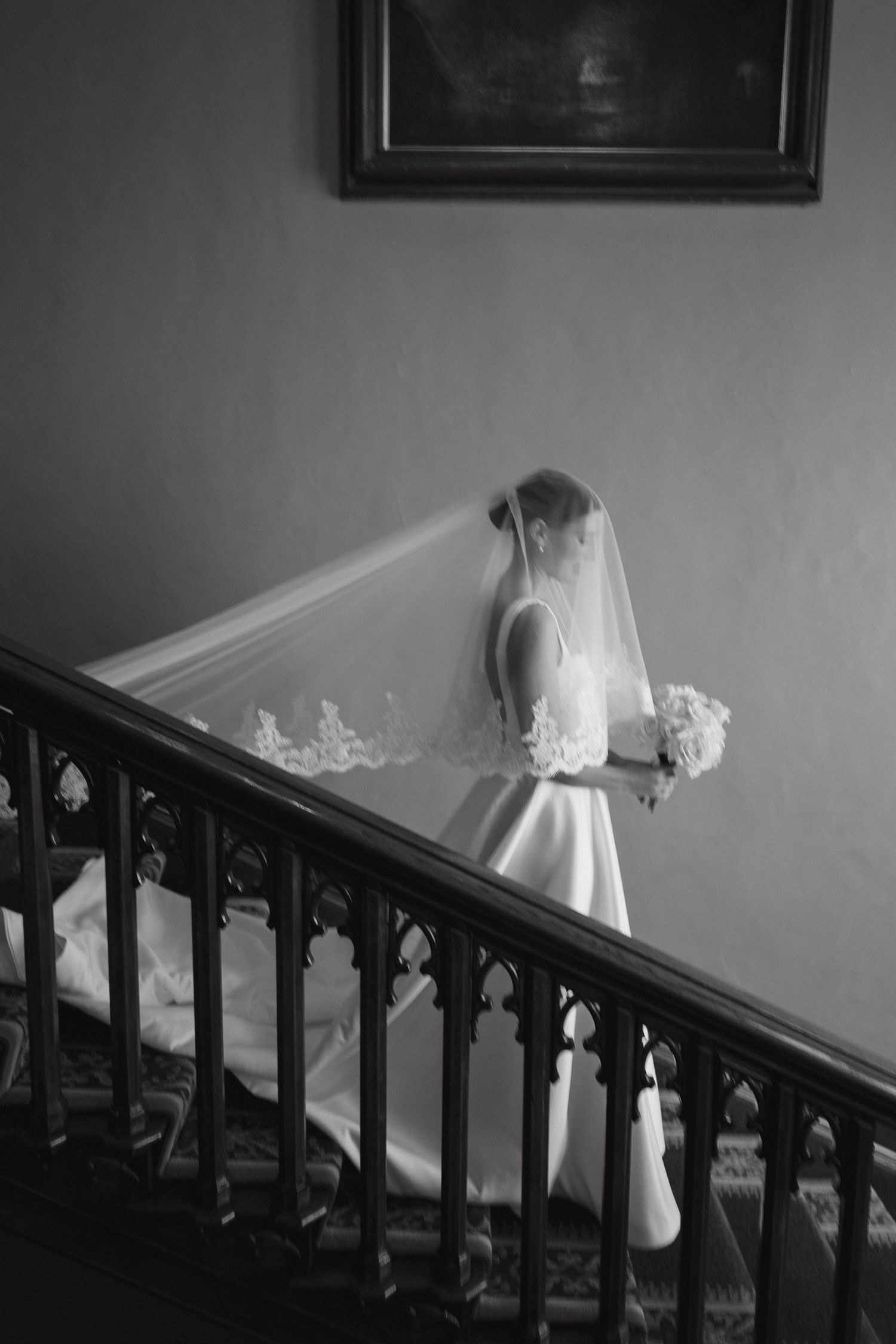 Black and white photo of bride in flowing dress descending ornate staircase.
