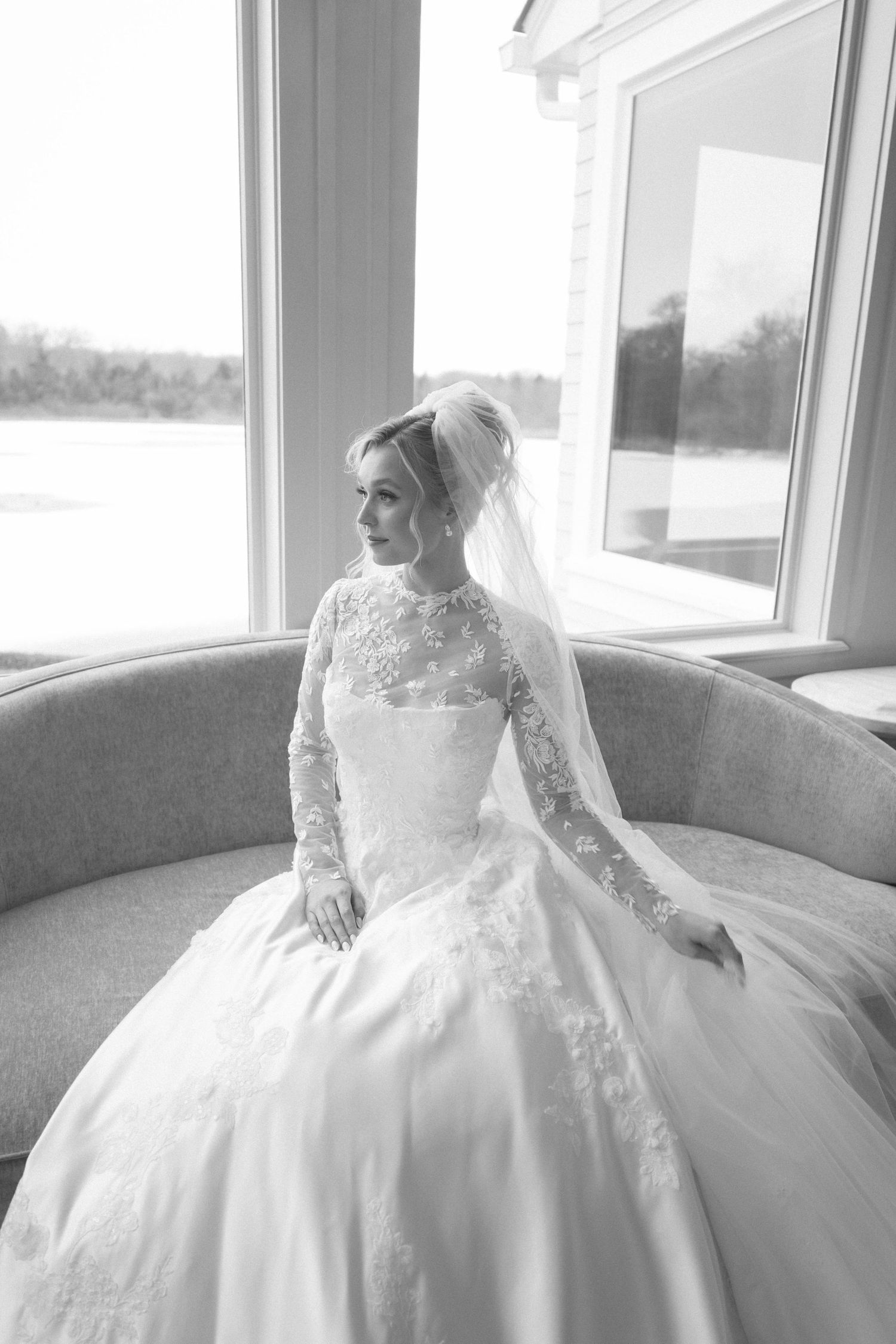 Bride in an elegant lace wedding gown poses by window in black and white portrait series.