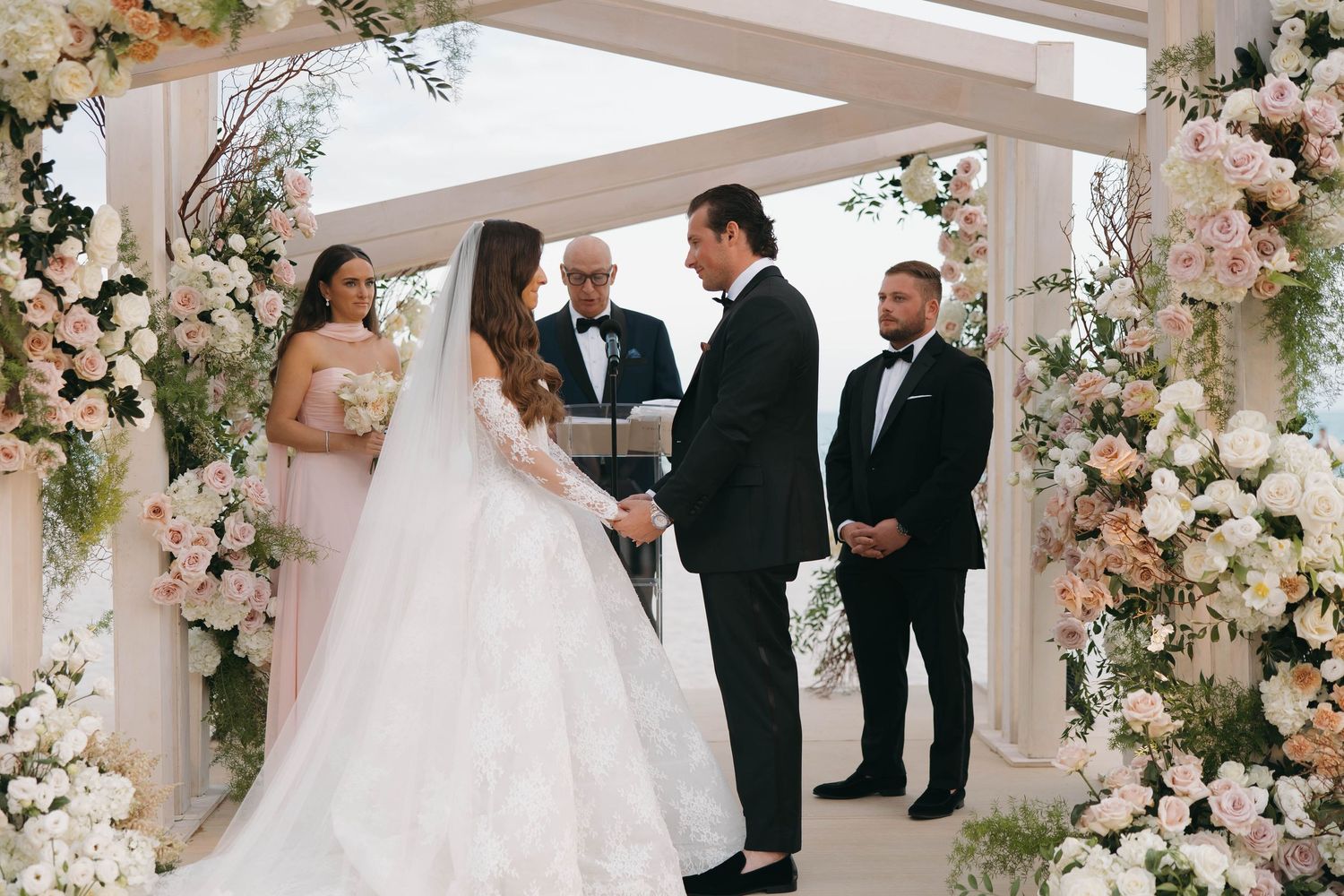 A wedding ceremony takes place under an elegant white arbor adorned with lush floral arrangements and greenery.