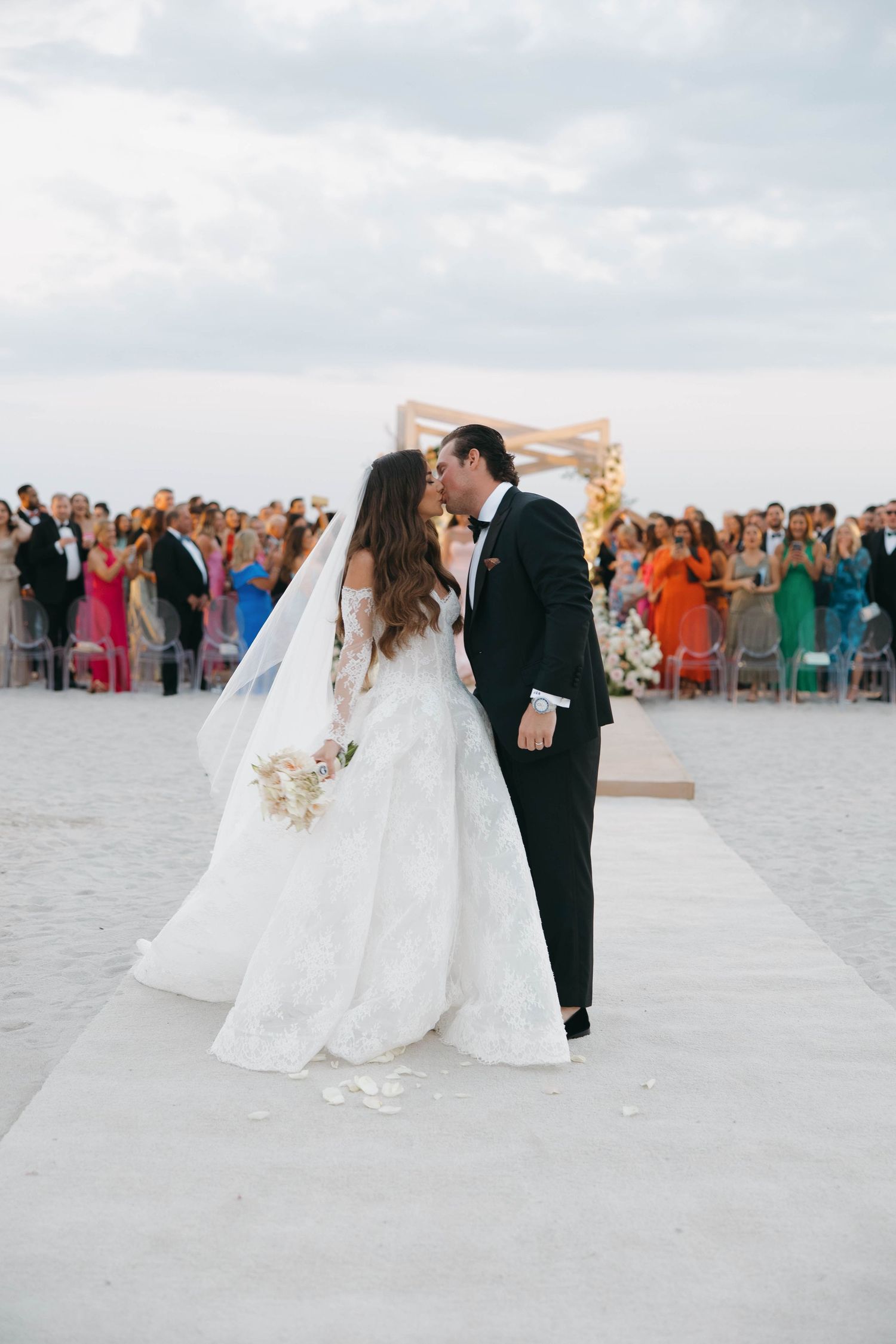 Wedding party celebrates on a white sandy beach with guests in colorful attire looking on.