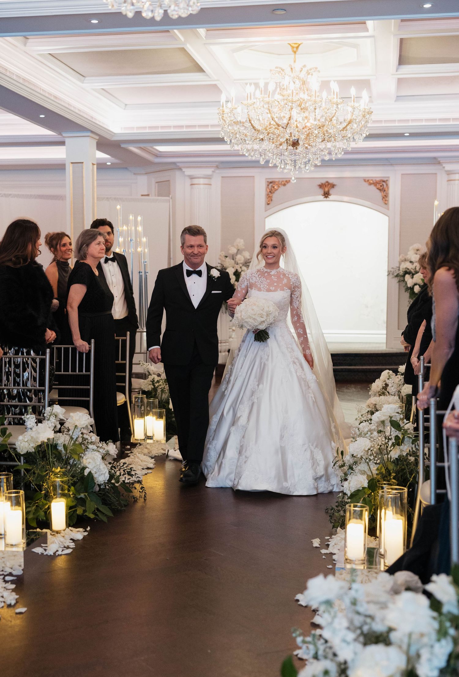 Elegant wedding ceremony setup with white floral arrangements and candlelit aisle in a grand ballroom.