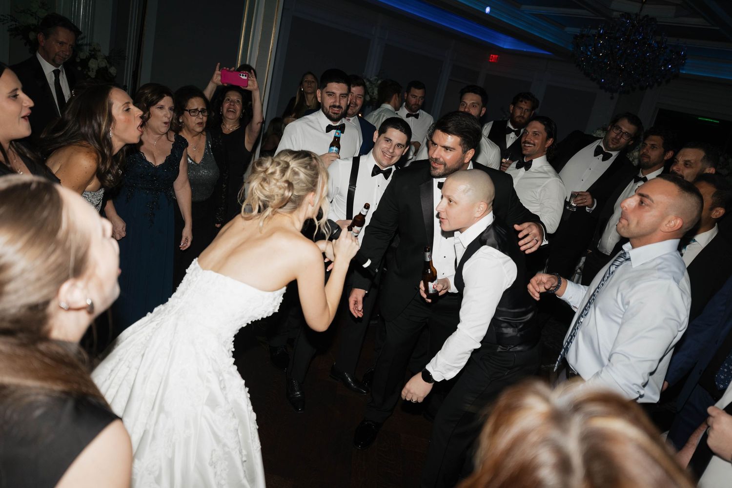 Wedding party dances energetically at reception with guests gathered around in formal attire.