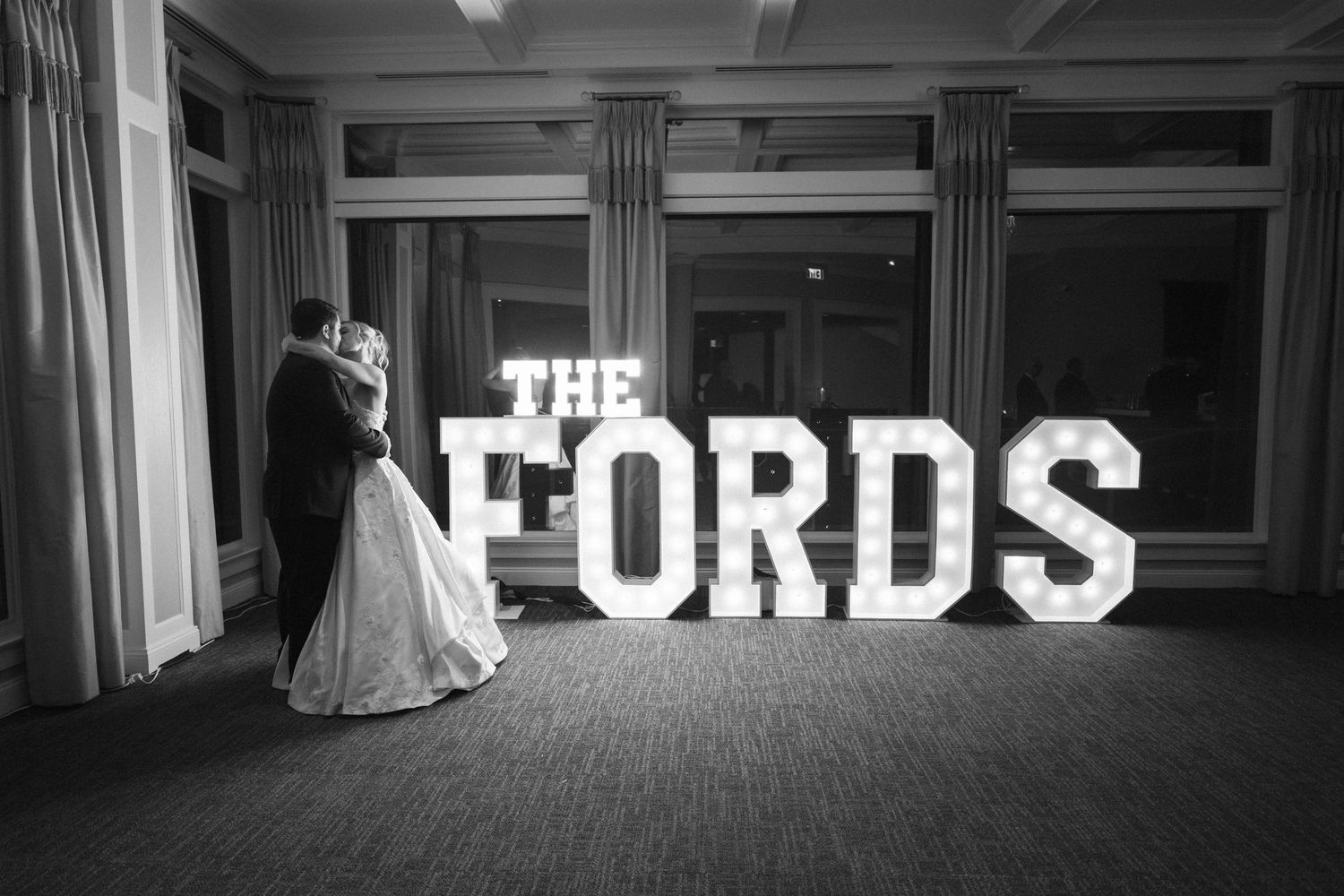 Black and white panoramic shot of a wedding reception with lit signage reading 'THE FORDS'.