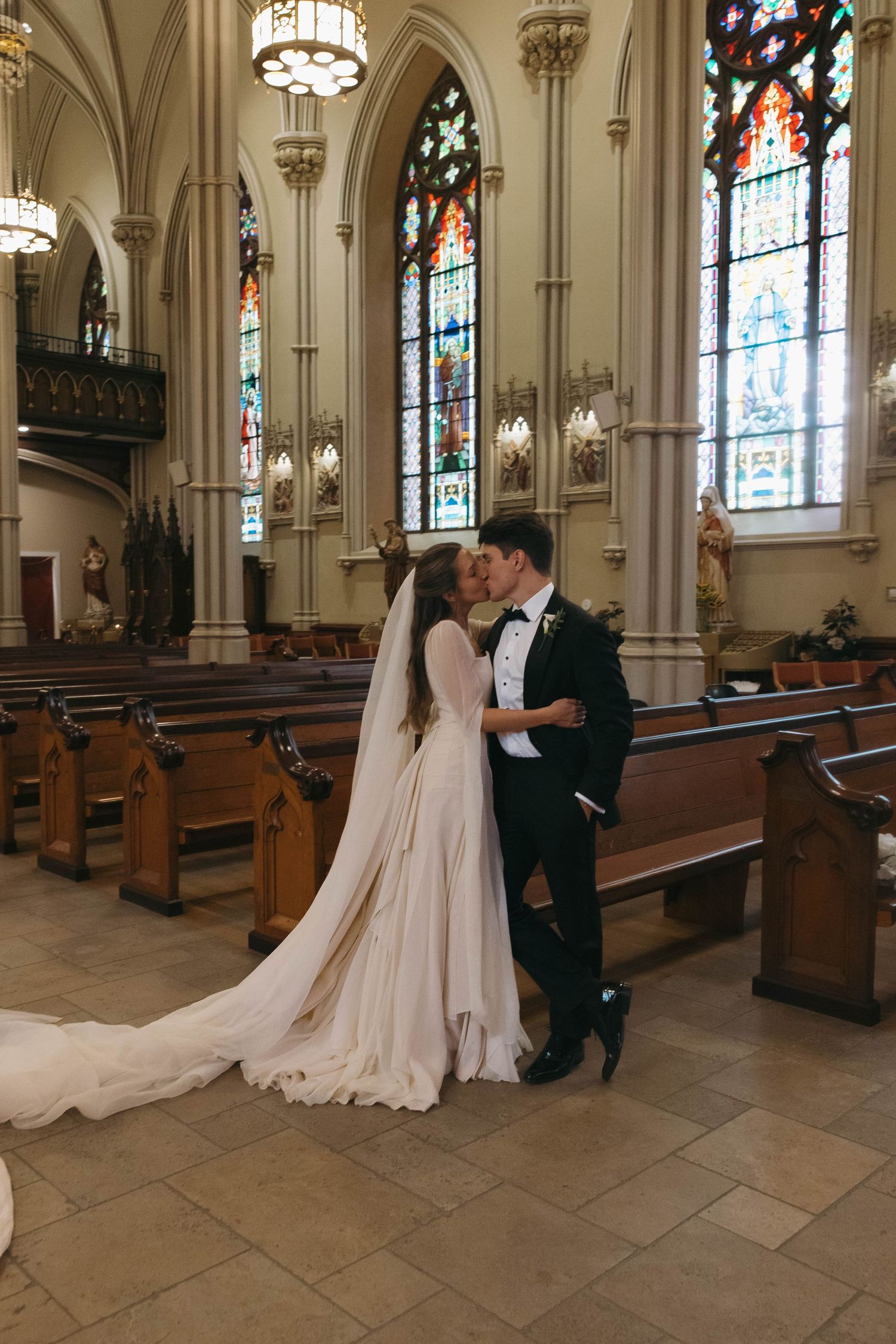 Bride and groom share first kiss in elegant church with stained glass windows and wooden pews on wedding day.