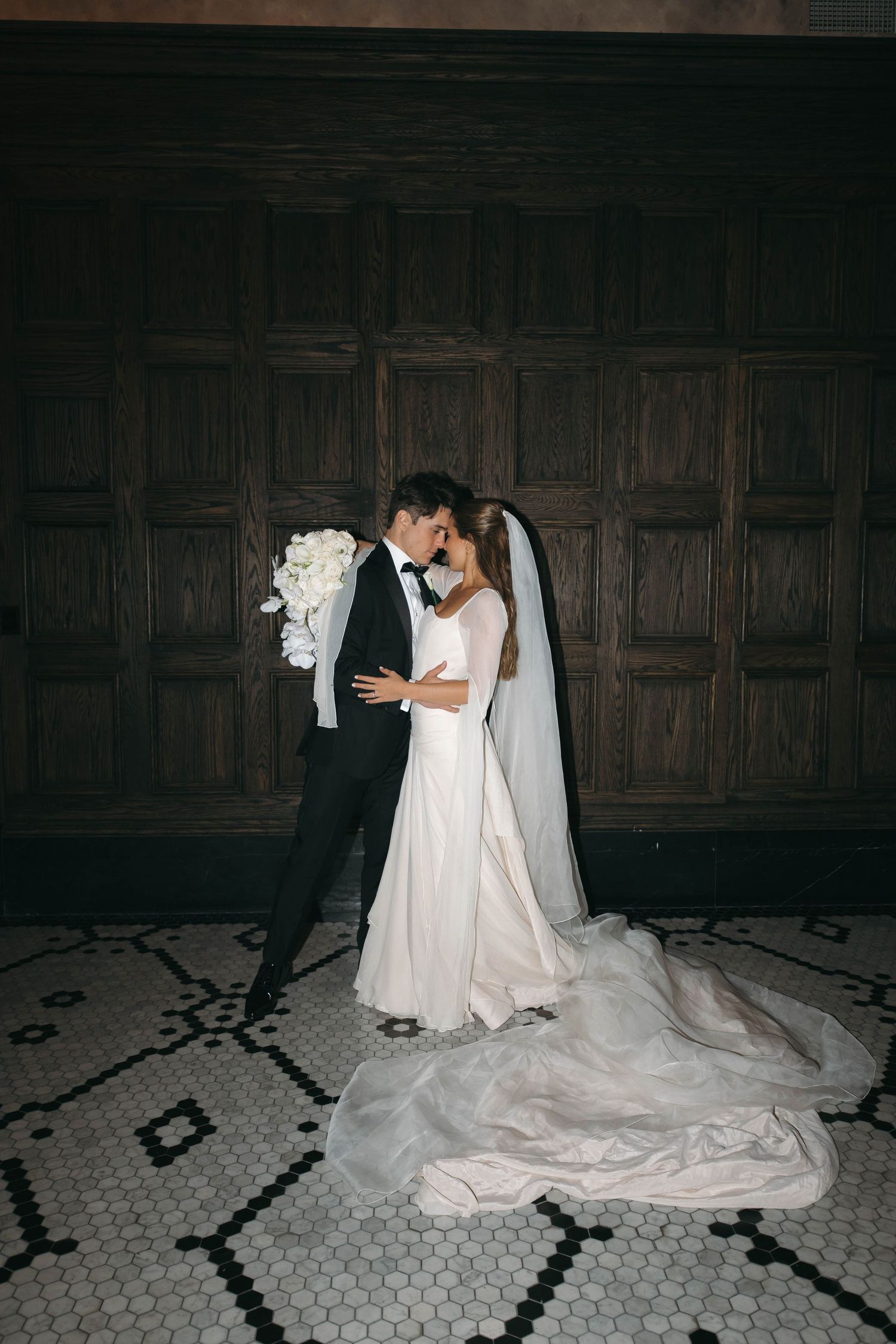 Elegant bride and groom in black tuxedo and white gown with veil embrace before ornate wooden doors on patterned tile floor.
