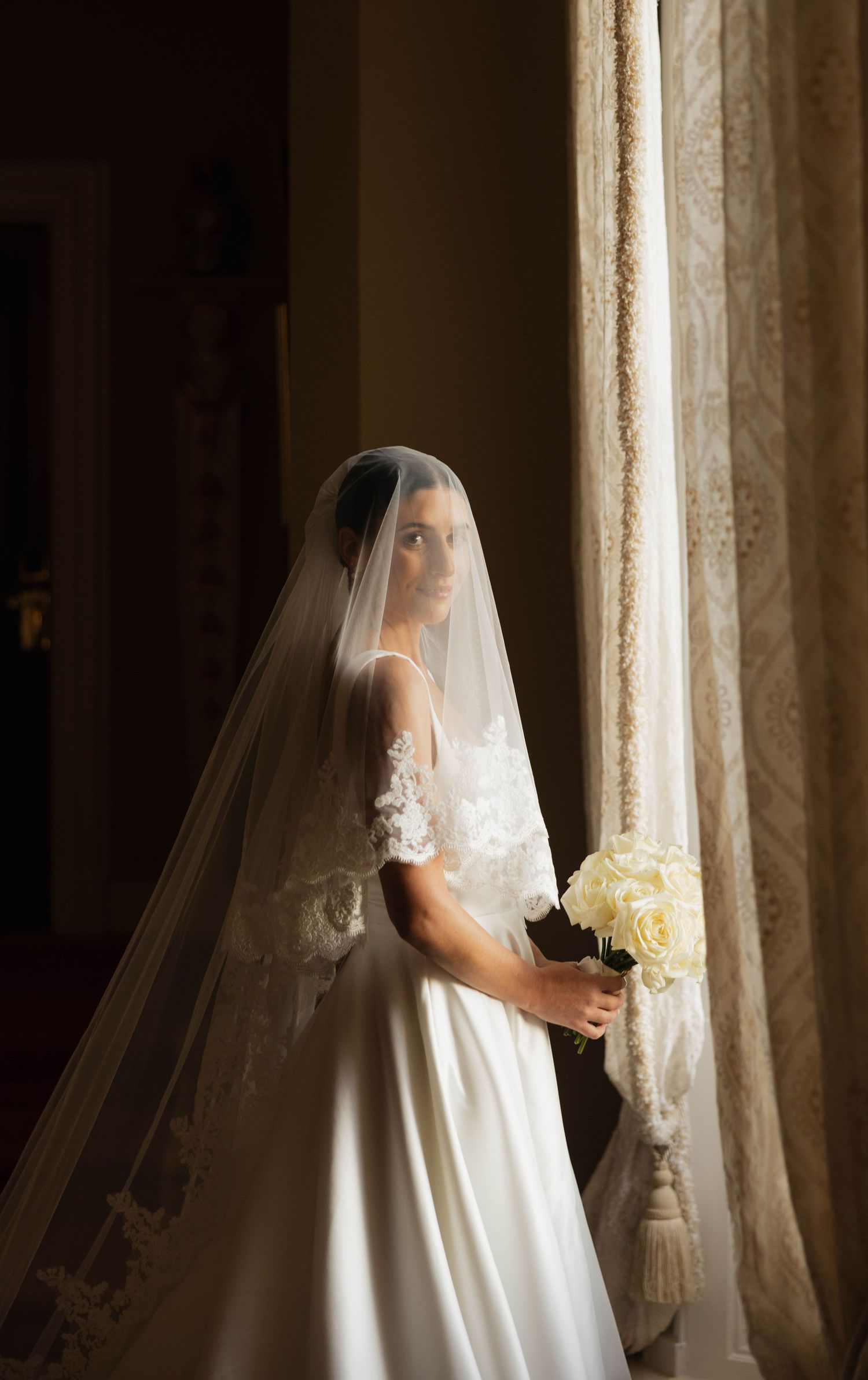 Ethereal bridal portrait by window with flowing veil and white bouquet.