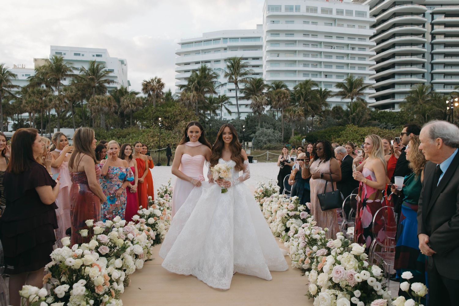 Beachfront wedding ceremony with white floral decorations and high-rise buildings in background.
