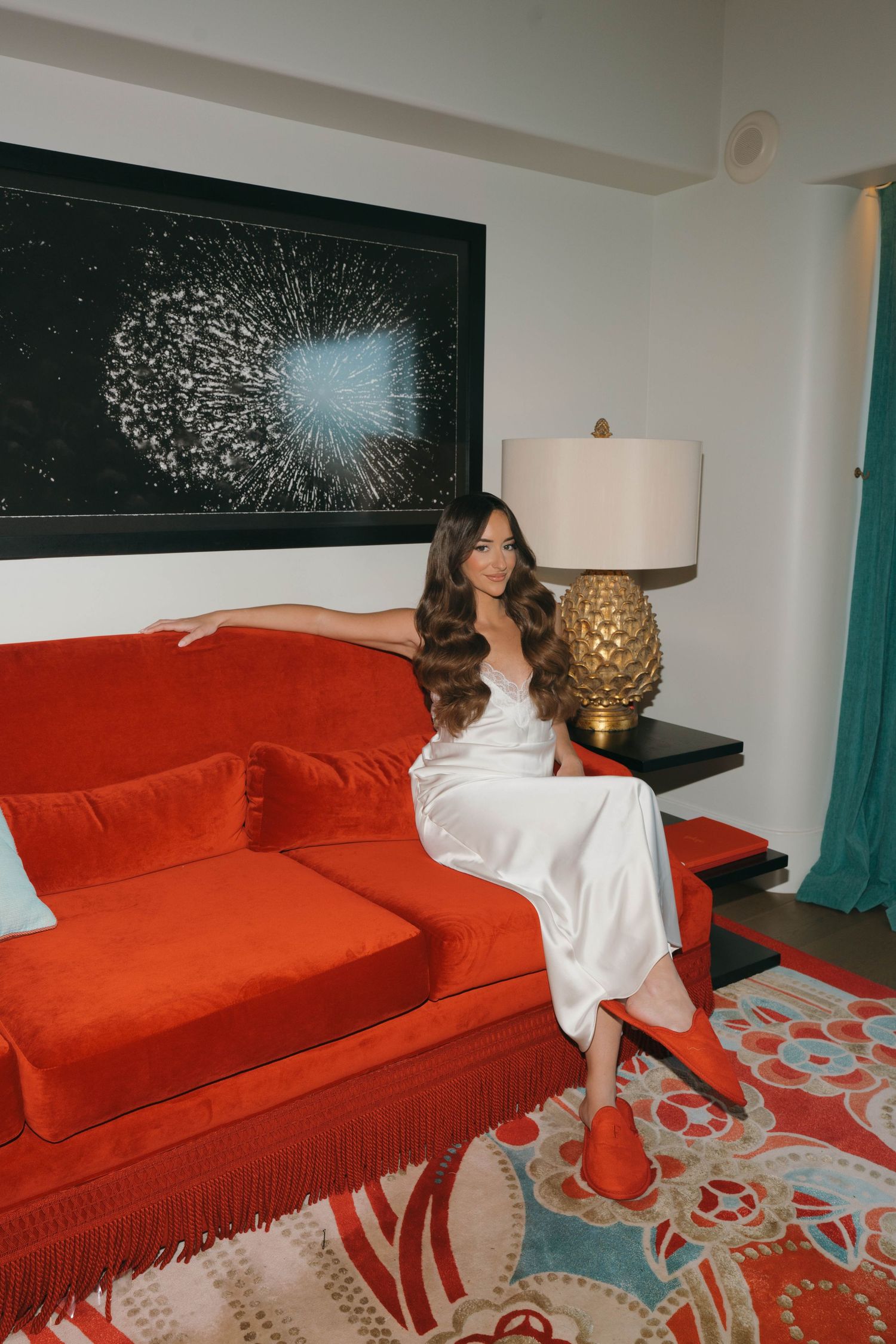 Person in white dress lounges on red couch with turquoise curtains and decorative artwork in background.