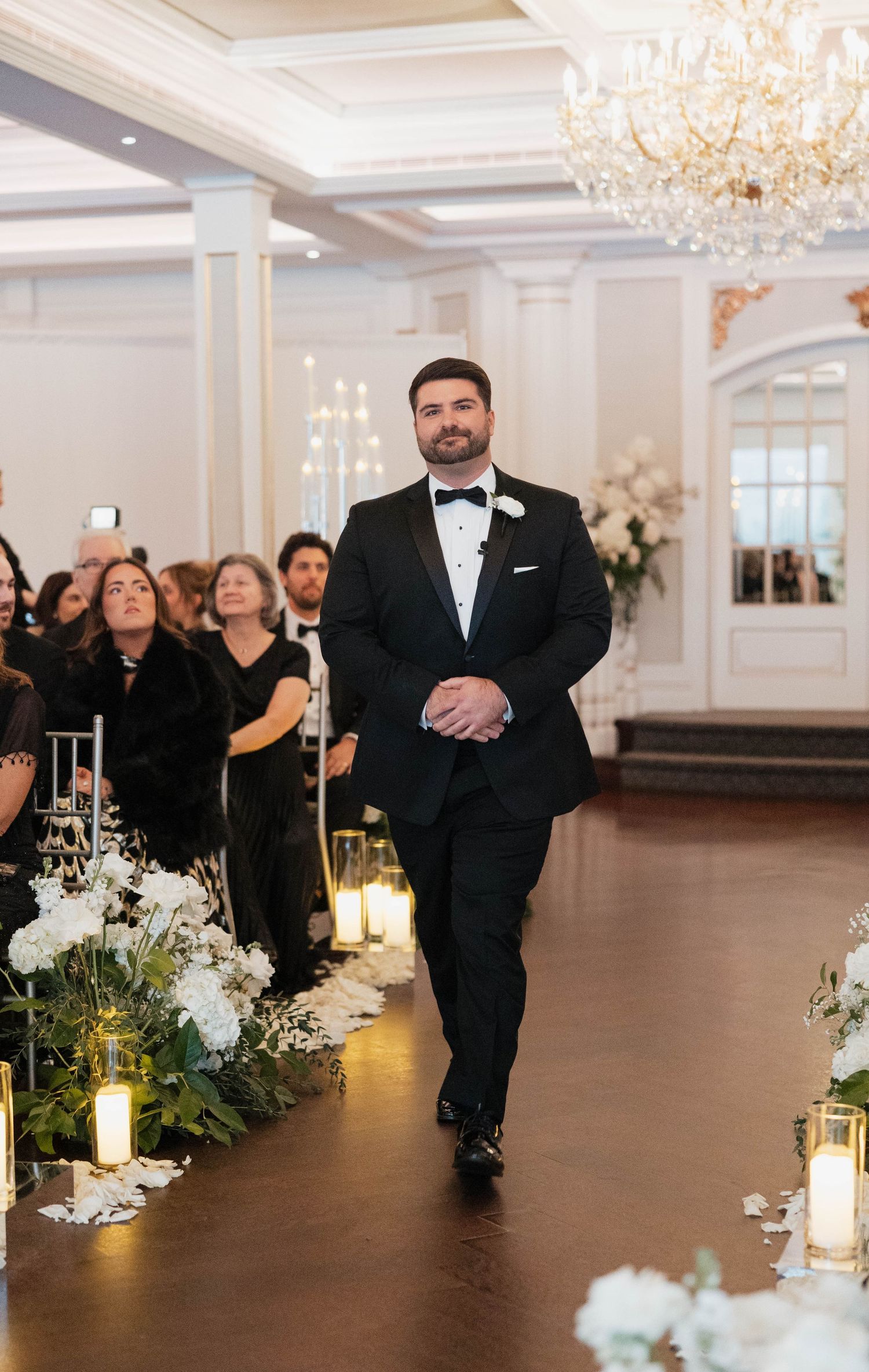 Wedding aisle lined with glowing candles and white floral arrangements in an elegant ballroom setting.
