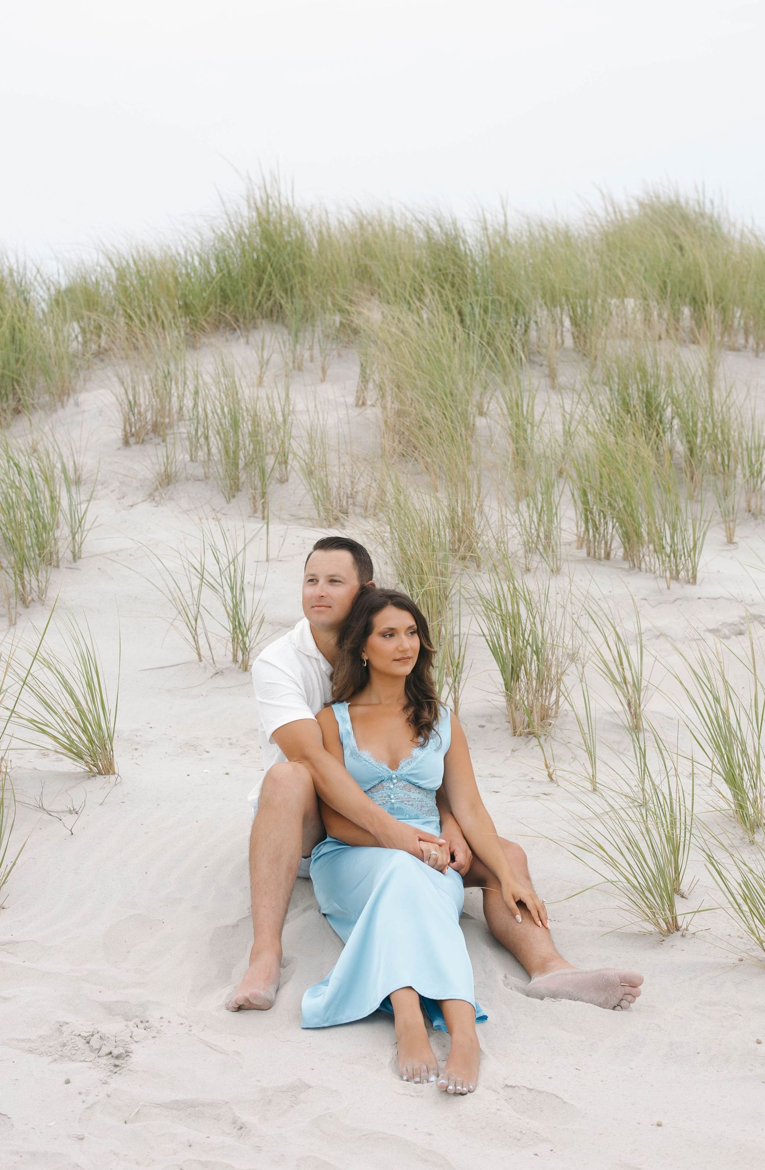 A couple in light blue and white outfits lounges on sand dunes with beach grass at sunset.
