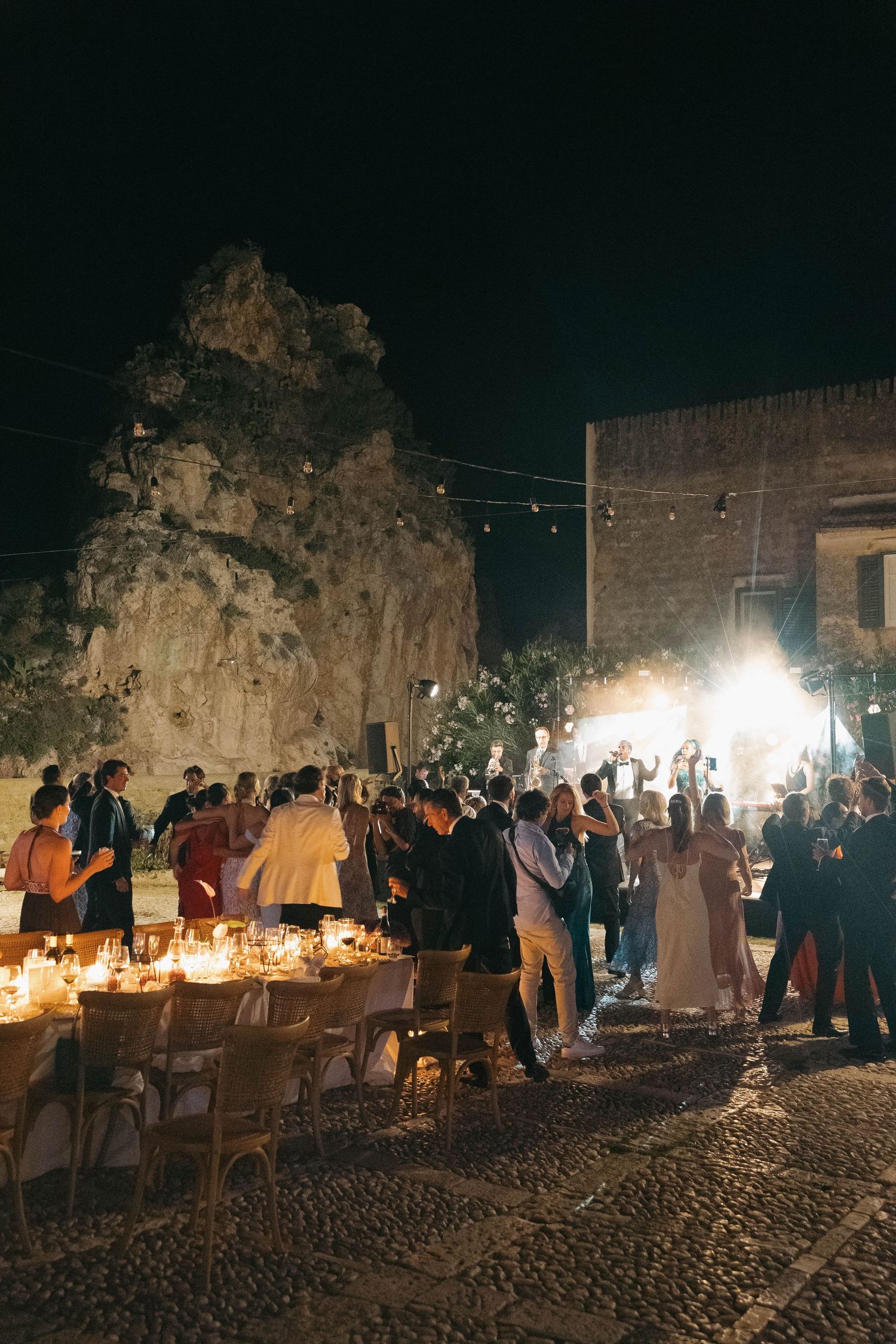 Festive outdoor gathering with candlelit tables against historic stone architecture.