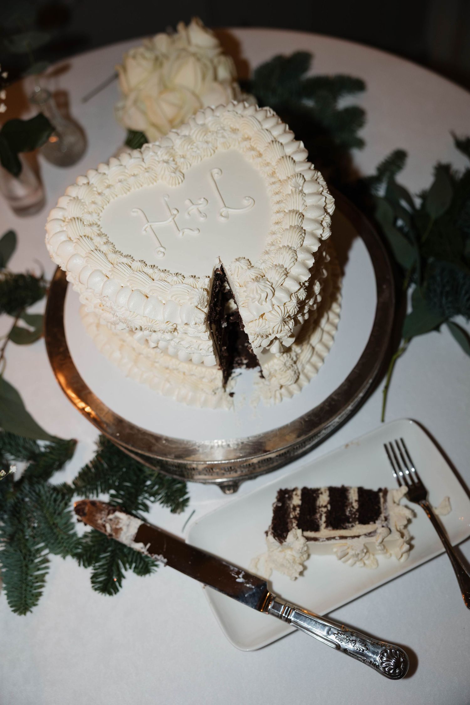 A white frosted layer cake decorated with piped details sits on a silver cake stand with greenery accents.