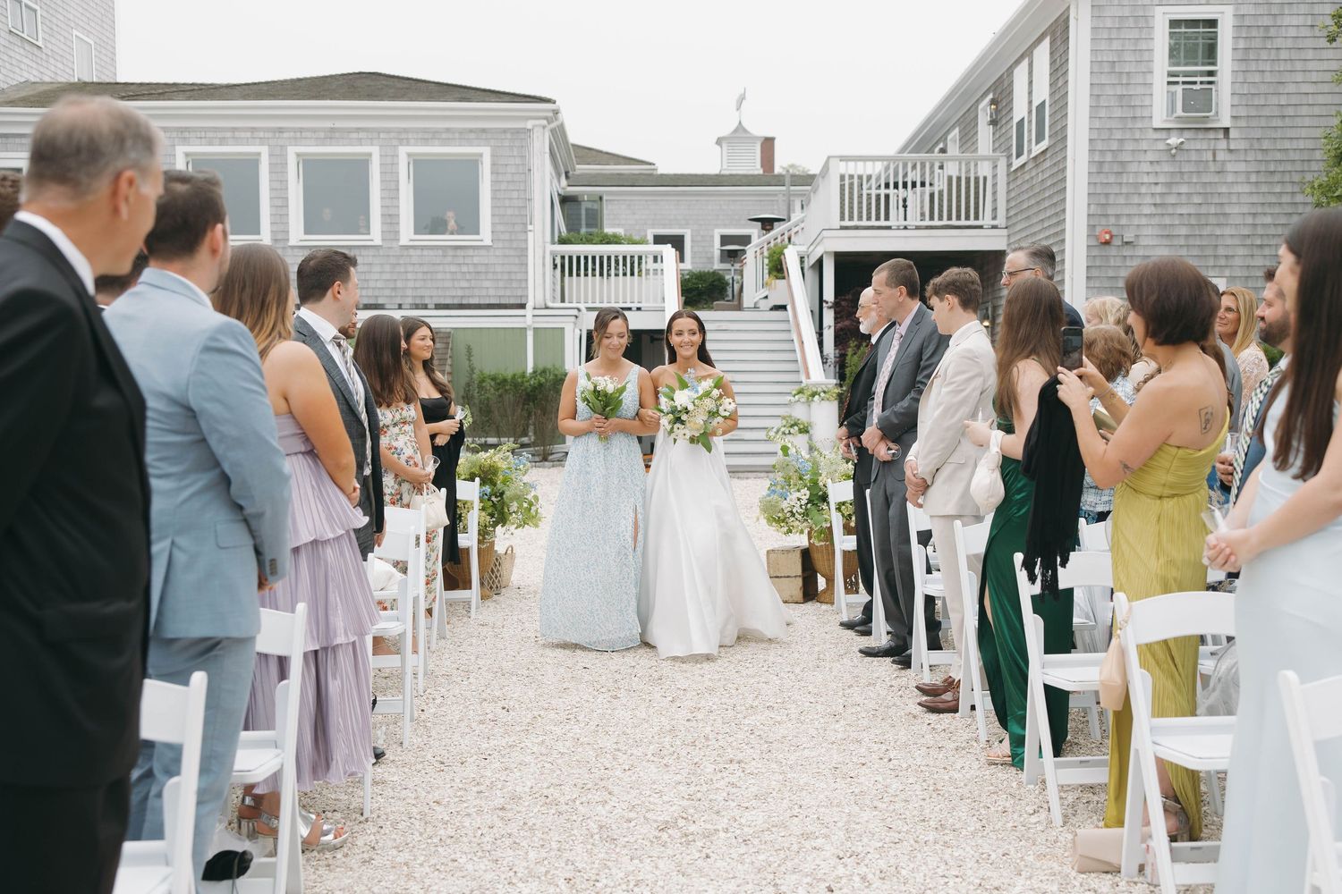 Wedding ceremony in progress on white gravel with coastal homes in background.