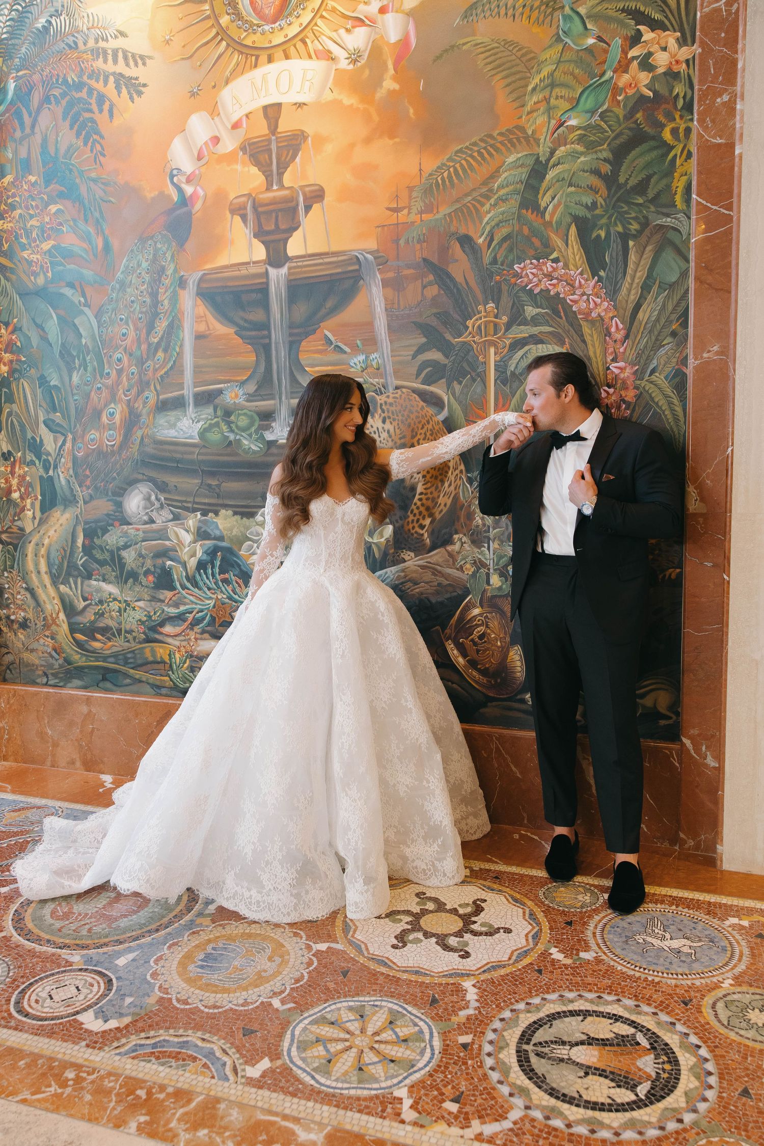 Bride in voluminous white gown dances with groom in front of decorative wall murals.