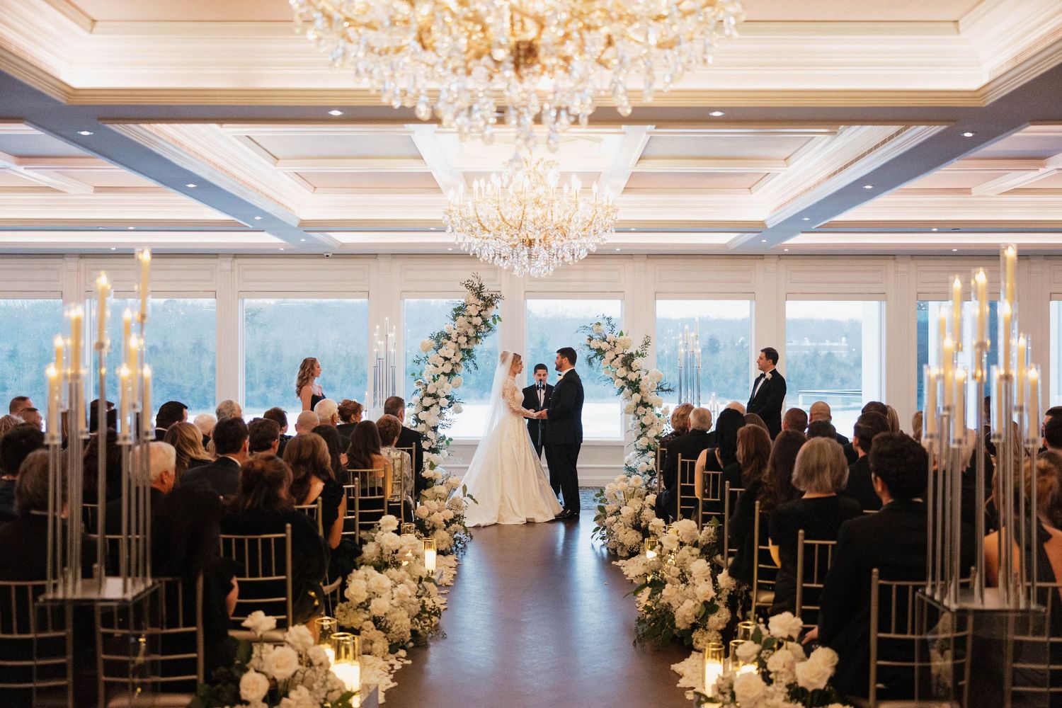 Luxurious reception hall with crystal chandeliers and white floral arrangements lining the aisle.