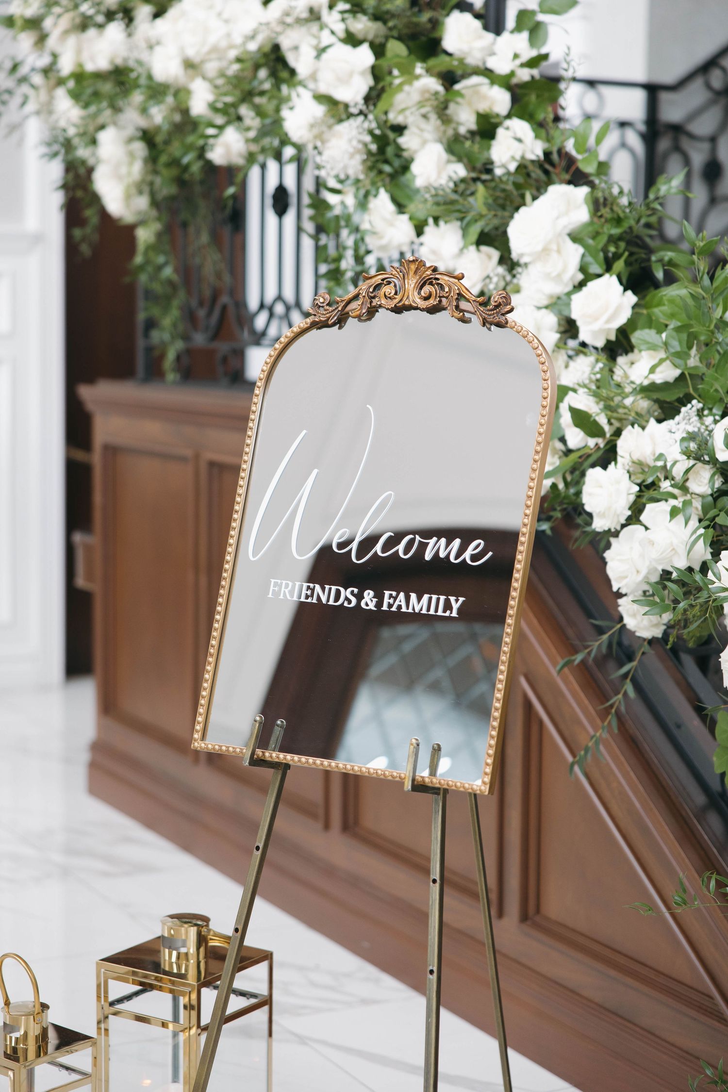 Ornate gold-framed mirror with 'Welcome Friends & Family' text surrounded by white floral arrangements.
