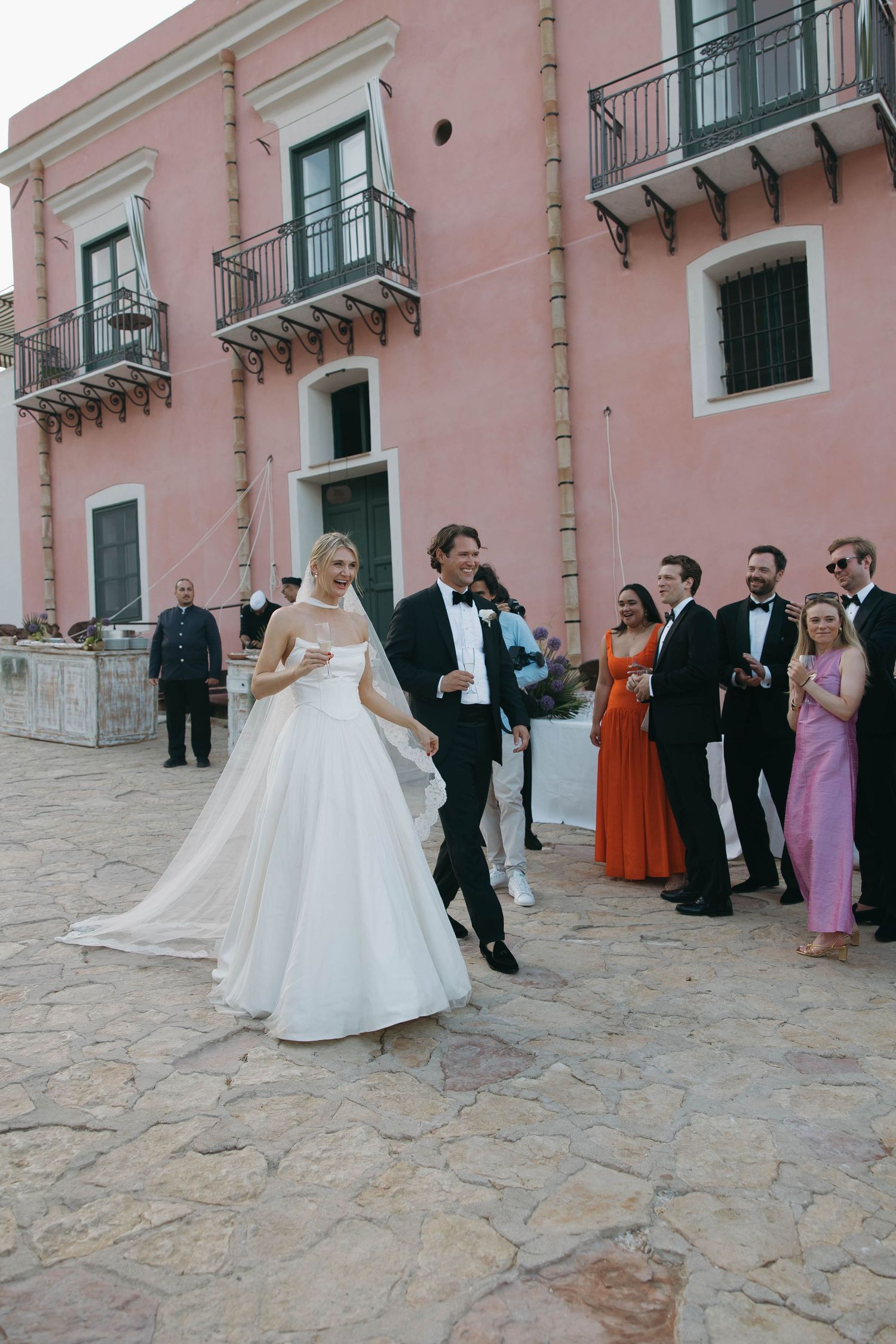 Elegant wedding celebration outside pink Mediterranean-style building with stone courtyard.