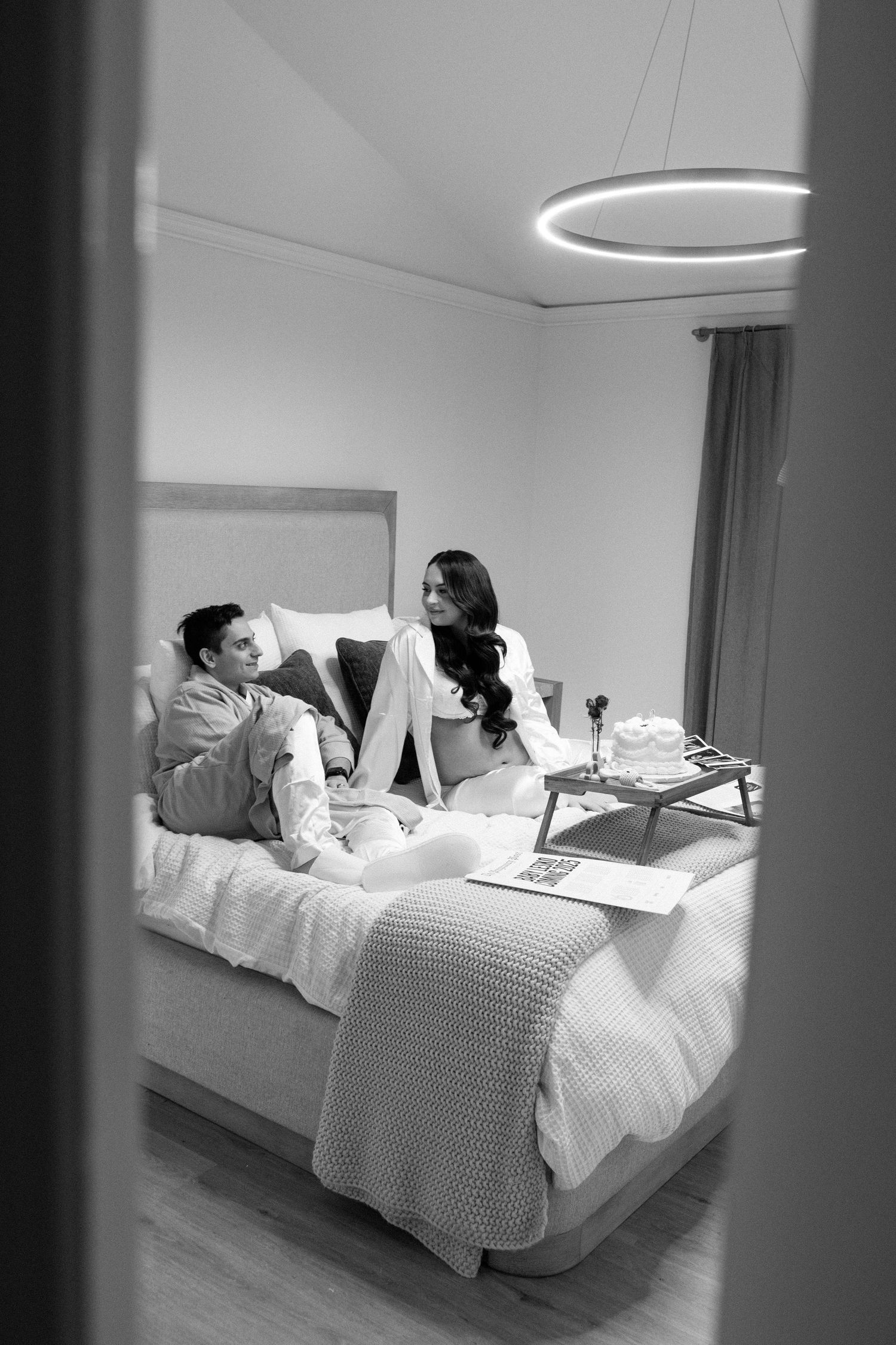 A couple relaxes on a bed with wine and snacks while having an intimate conversation in black and white.