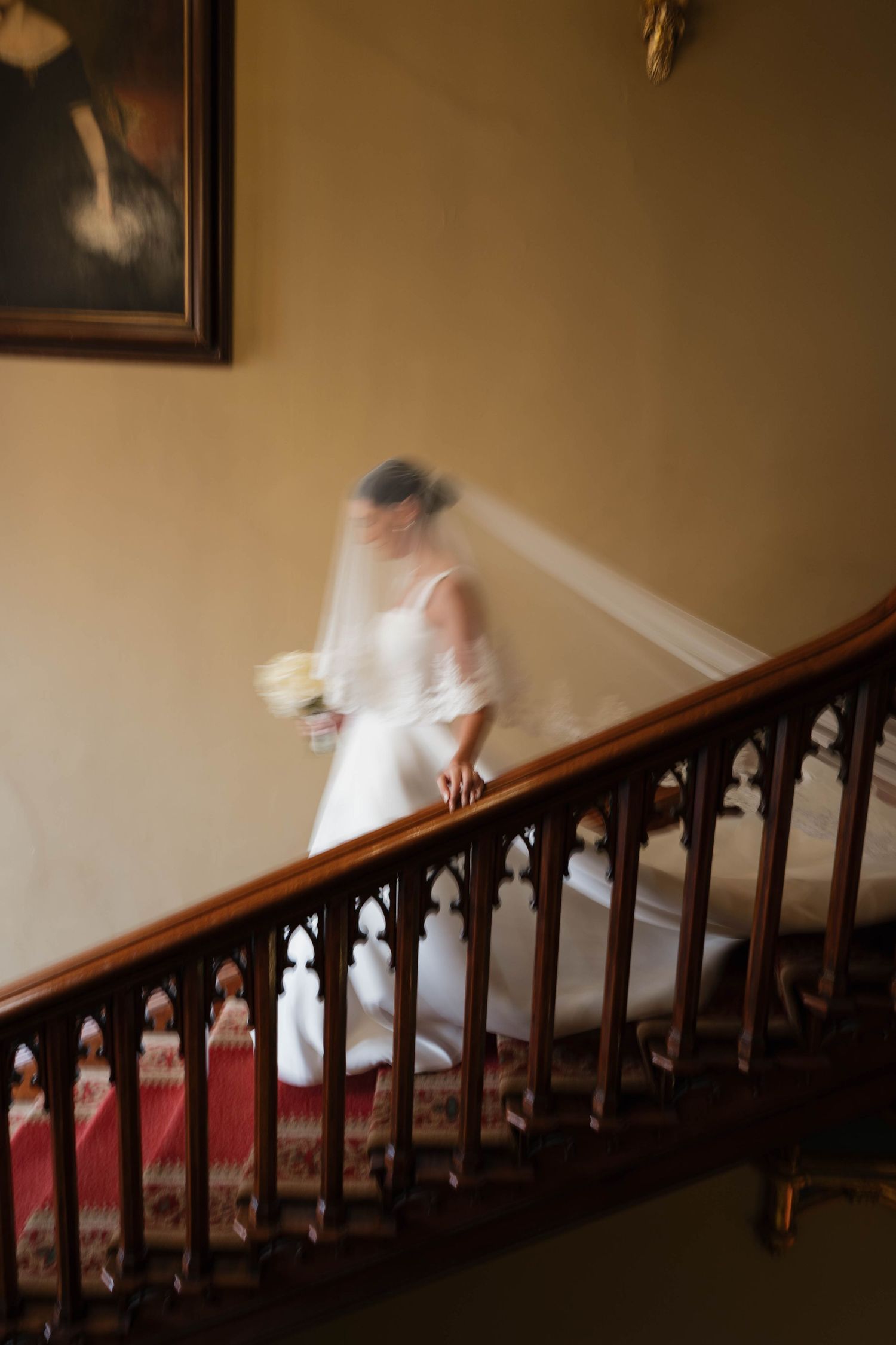 Motion-blurred image of bride descending ornate wooden staircase in vintage setting.