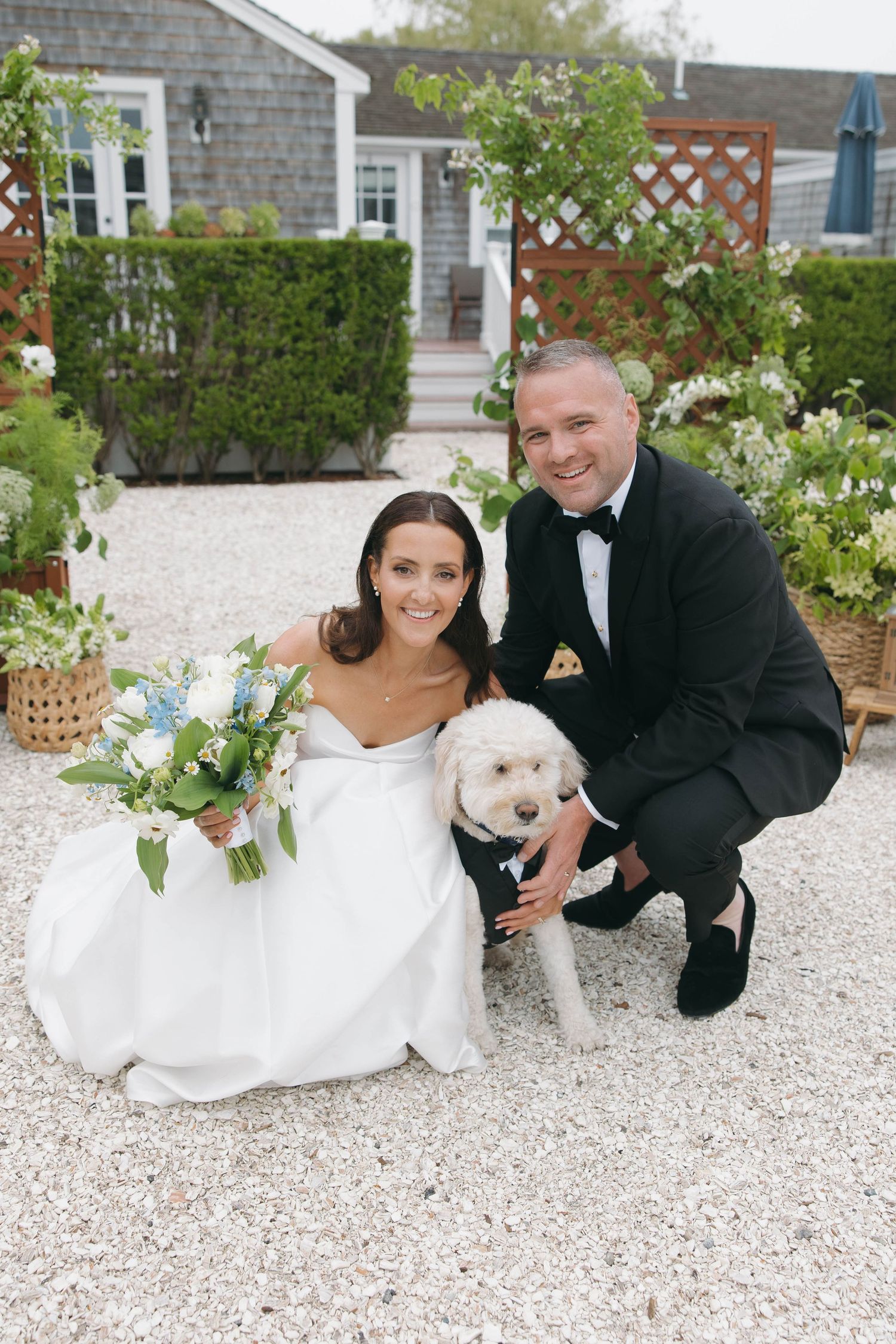 Wedding couple sits on gravel with their white dog in front of a shingle house.