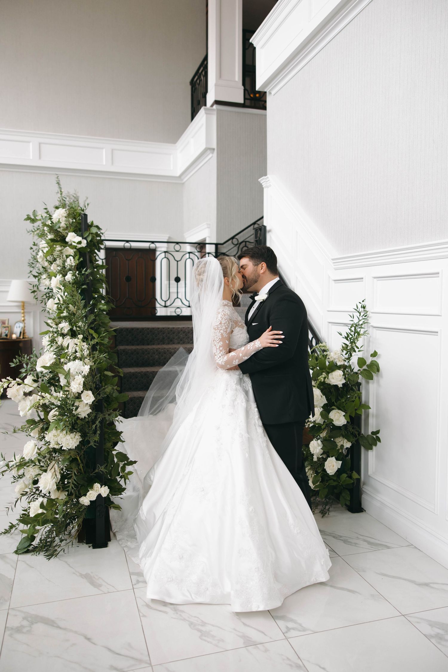 Bride and groom share intimate moments during first look with floral decorations in white hallway.