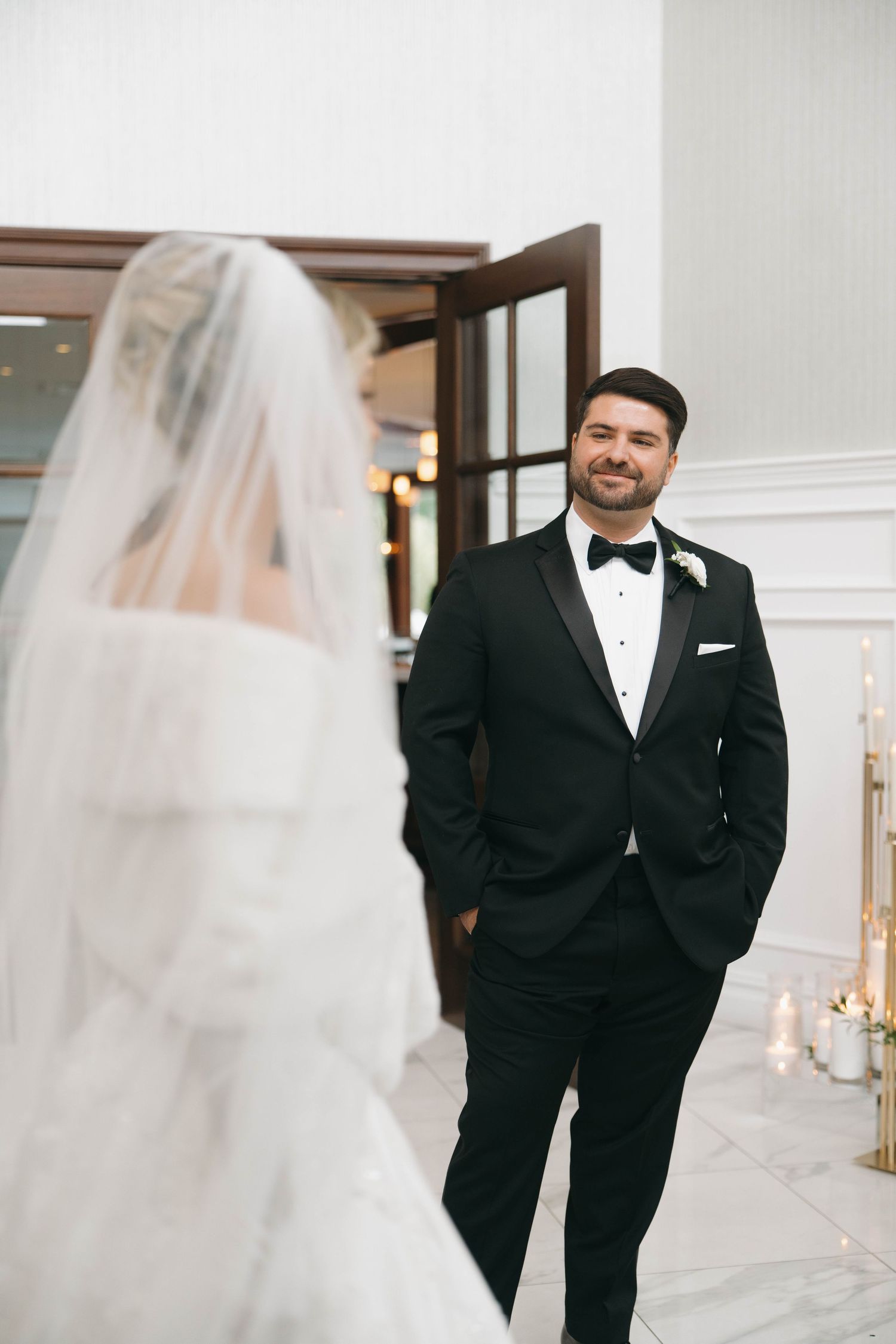 A groom in classic black tuxedo smiles during the first look wedding moment.