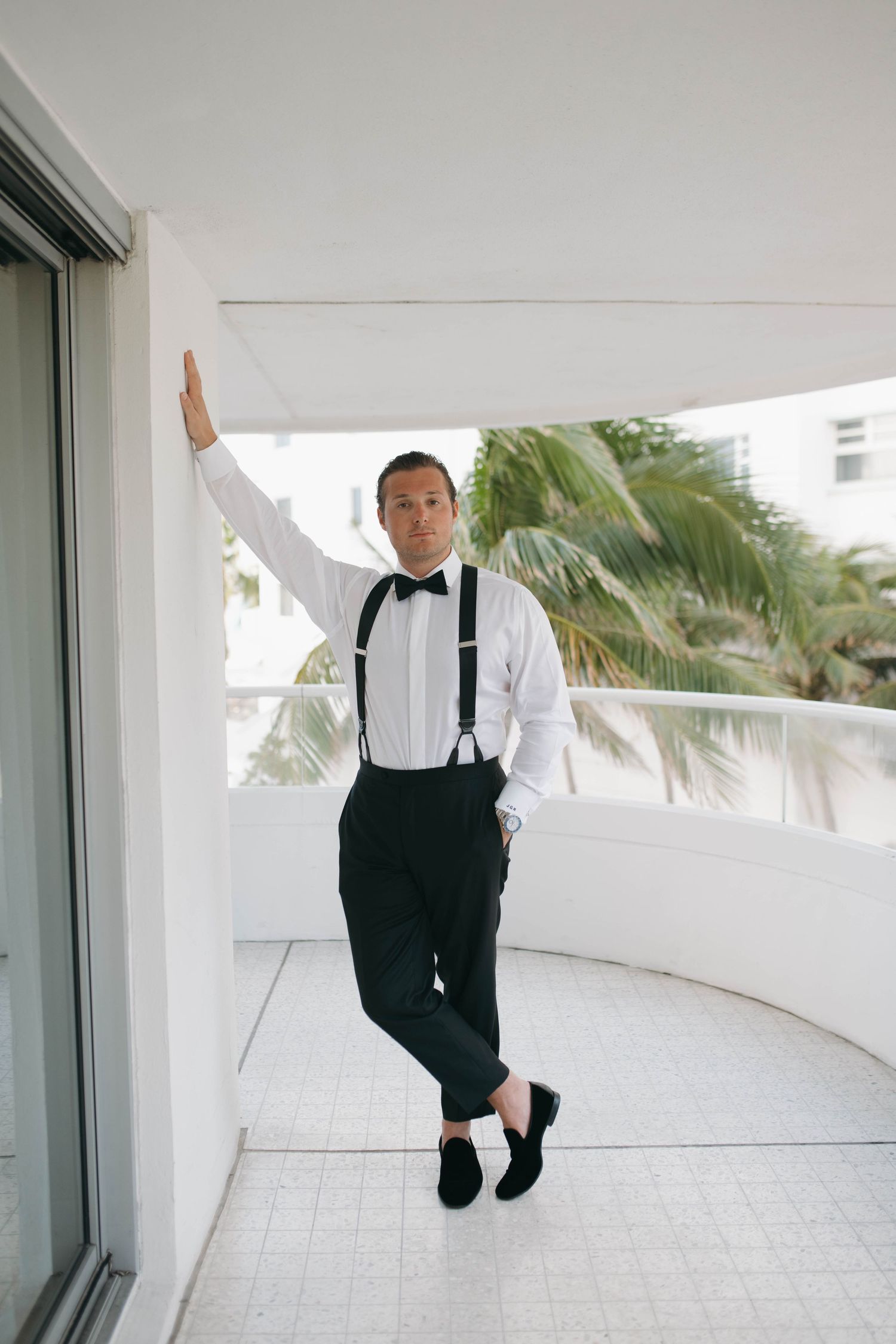 Person in formal wear jumping playfully on a white balcony with palm trees visible.