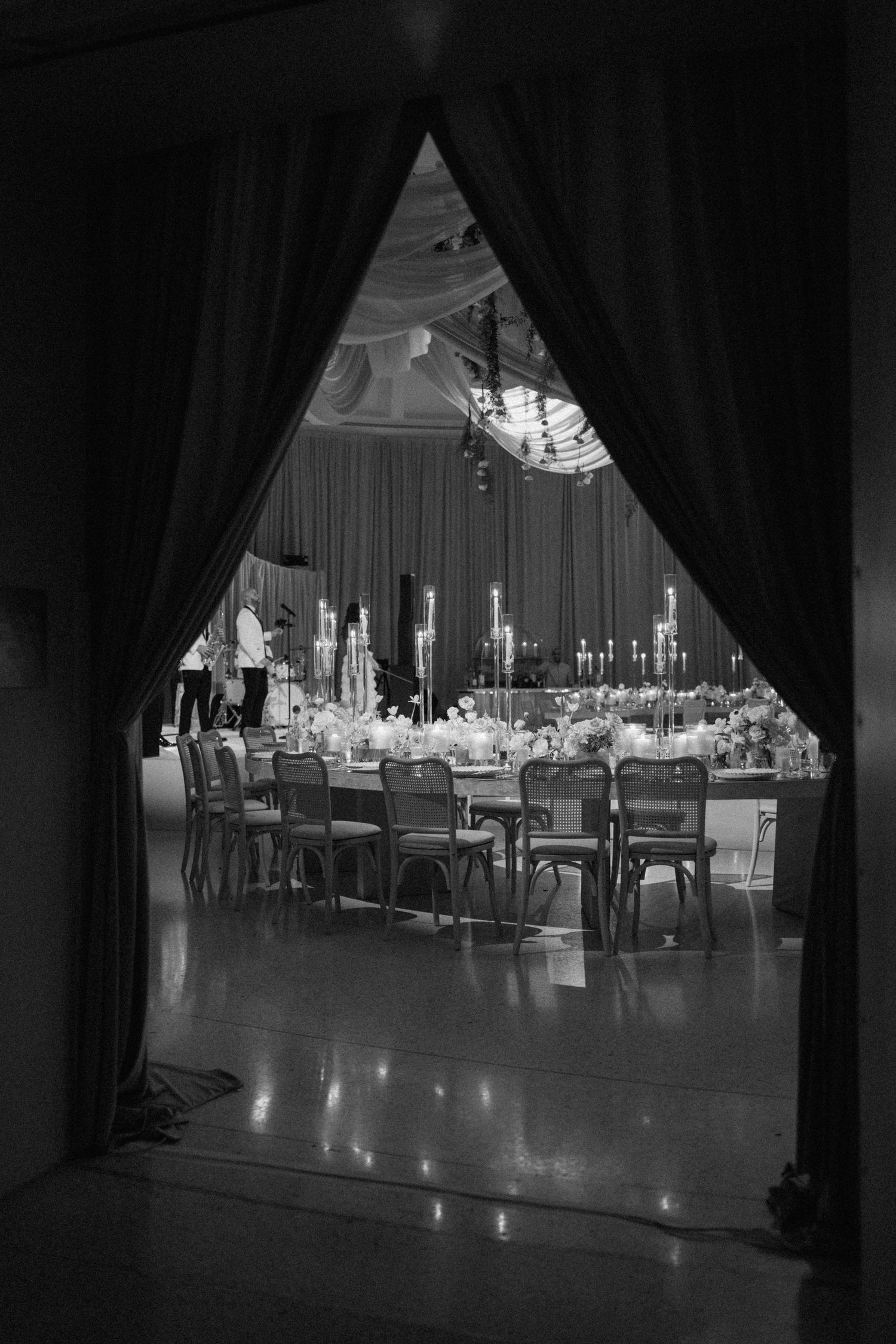 Black and white view of a wedding reception hall through parted curtains showing elegant table settings.
