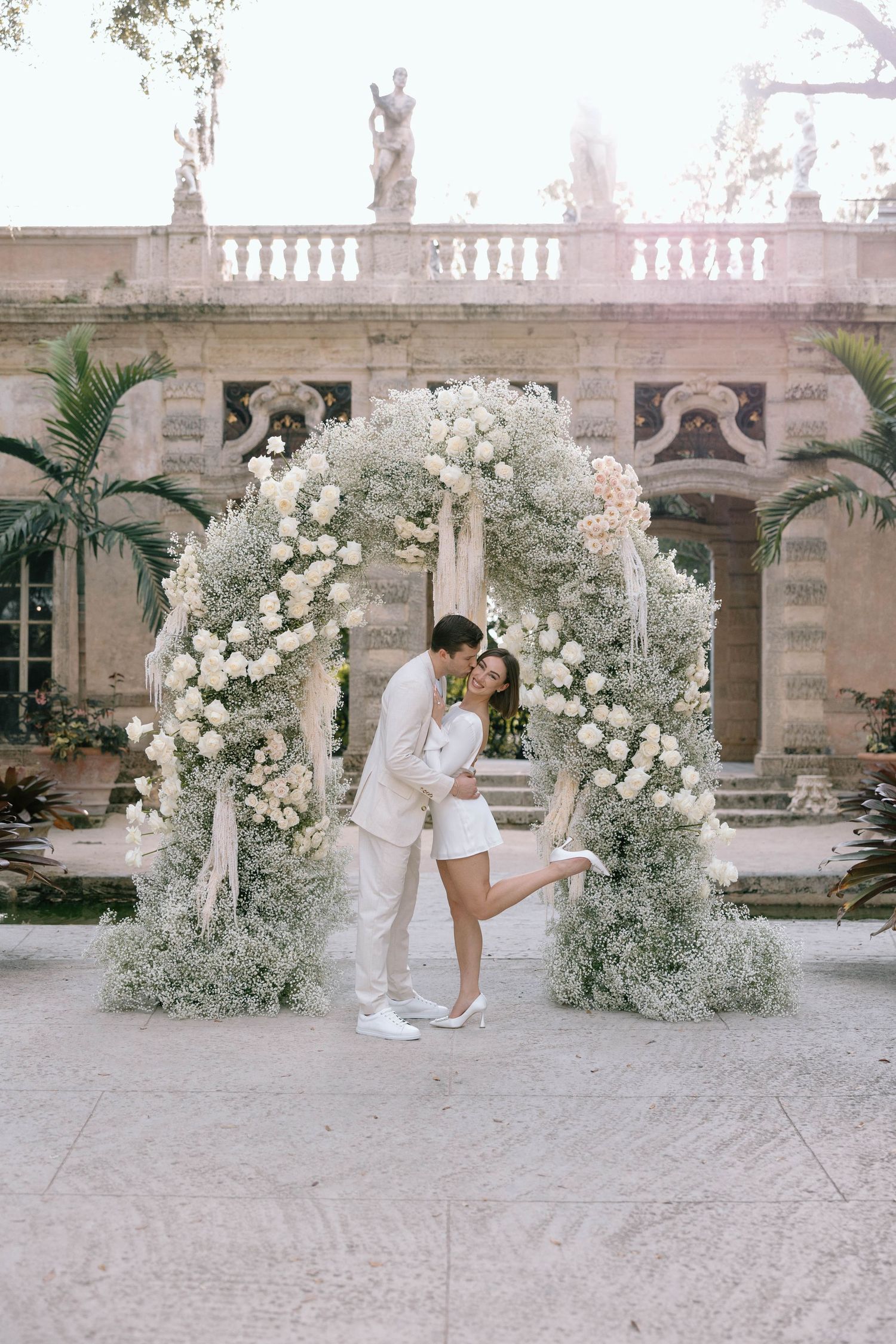 A couple shares a romantic moment under white floral arches in an elegant courtyard.
