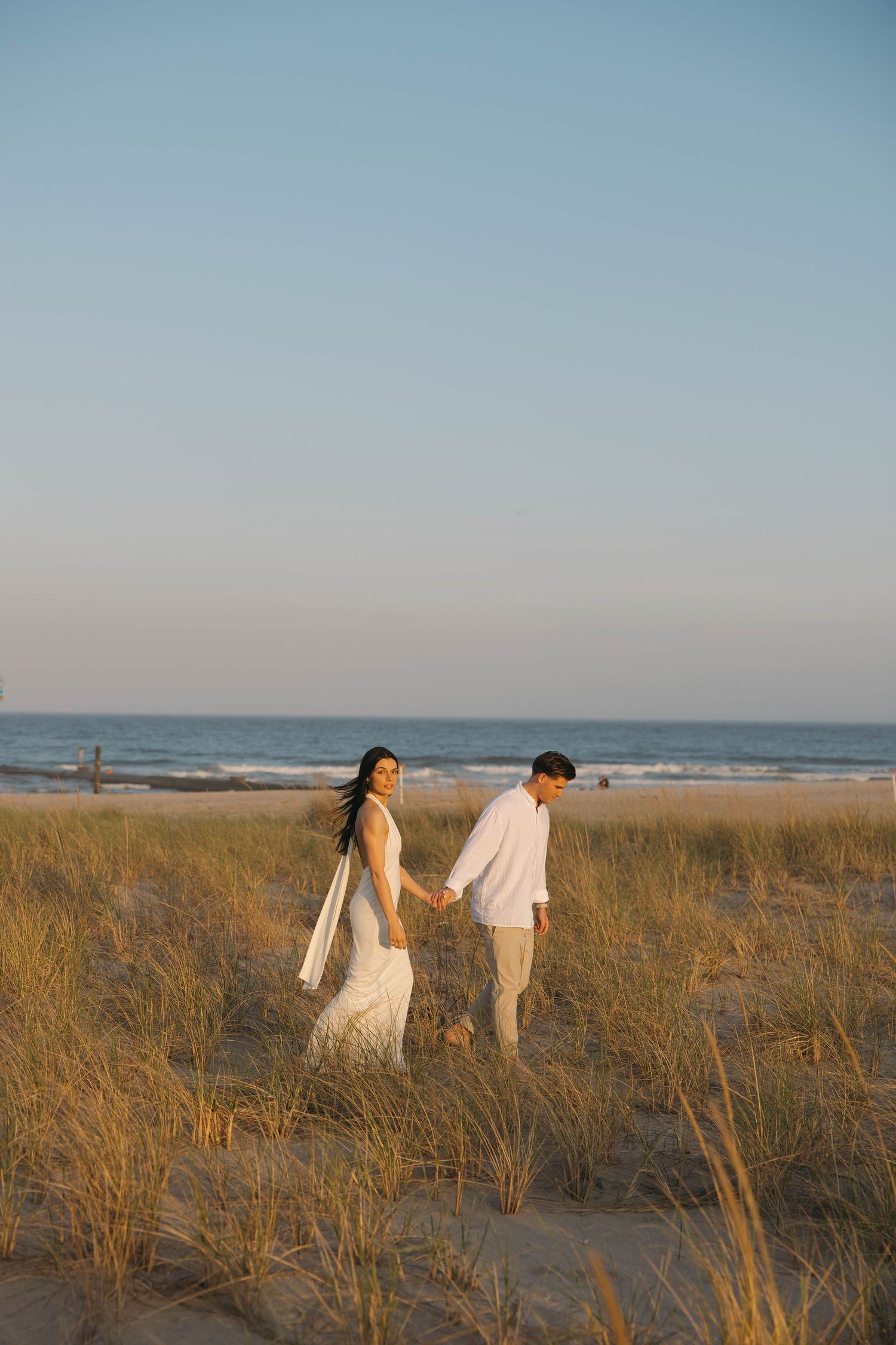 Couple walking hand in hand through coastal dunes at sunset.