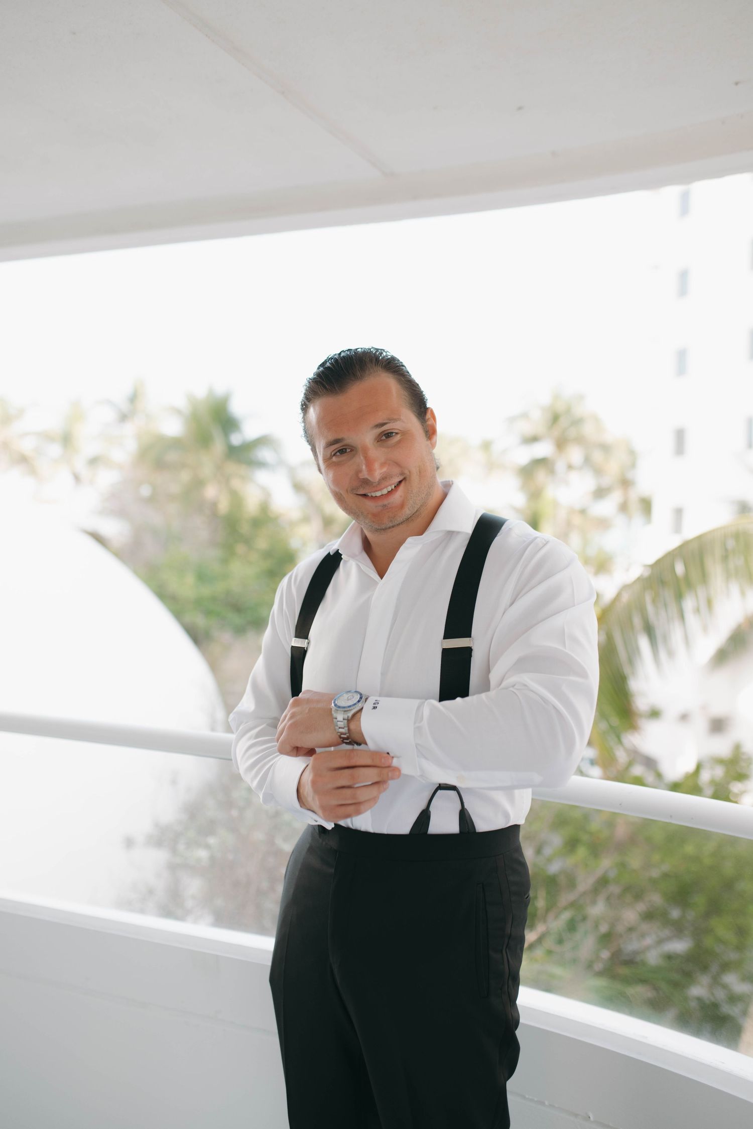 Person in formal attire with suspenders checking time on balcony with palm trees in background.