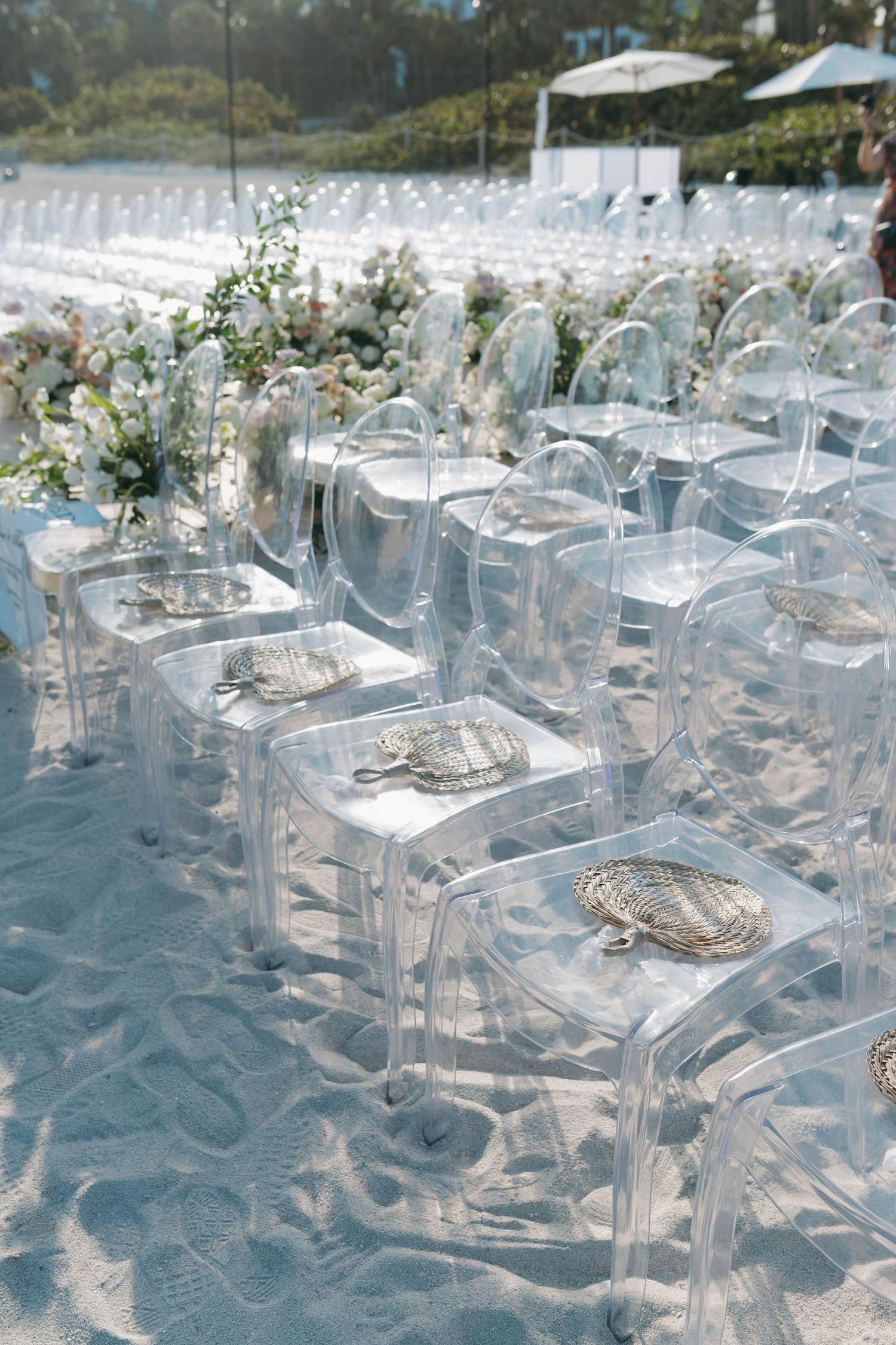 Ghost chairs arranged in rows for beach wedding ceremony with floral decorations.