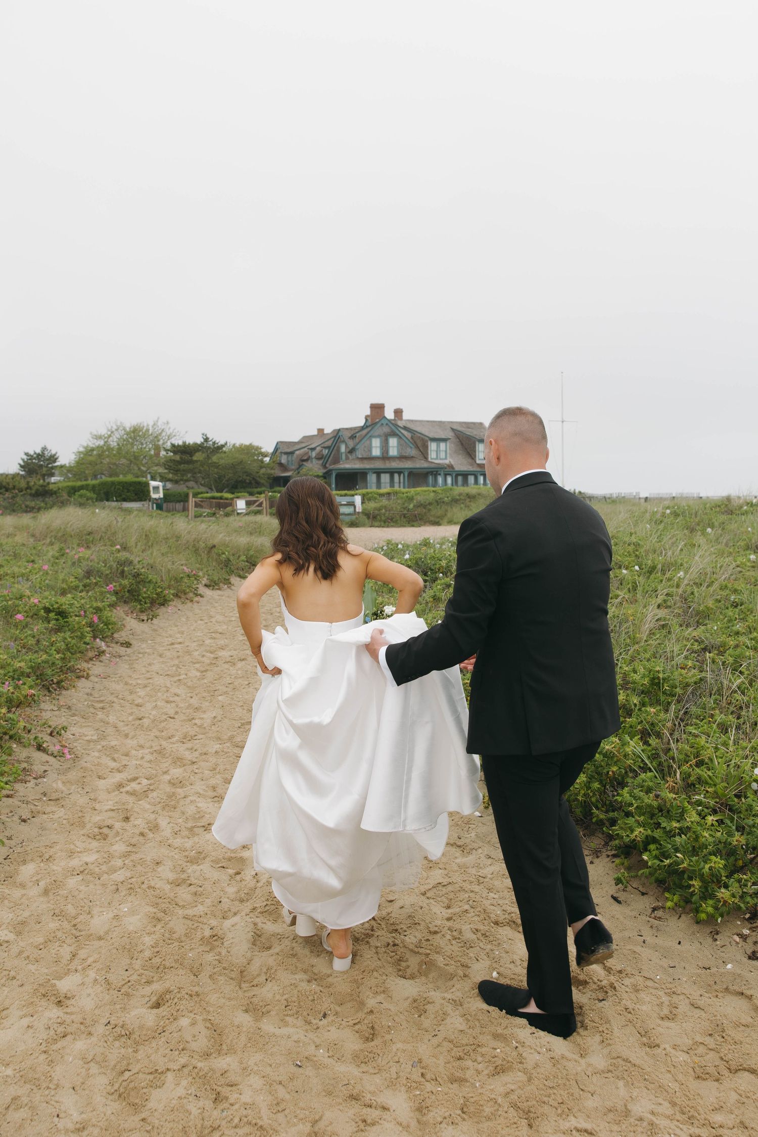 Couple walking up sandy path through beach grass on cloudy day.