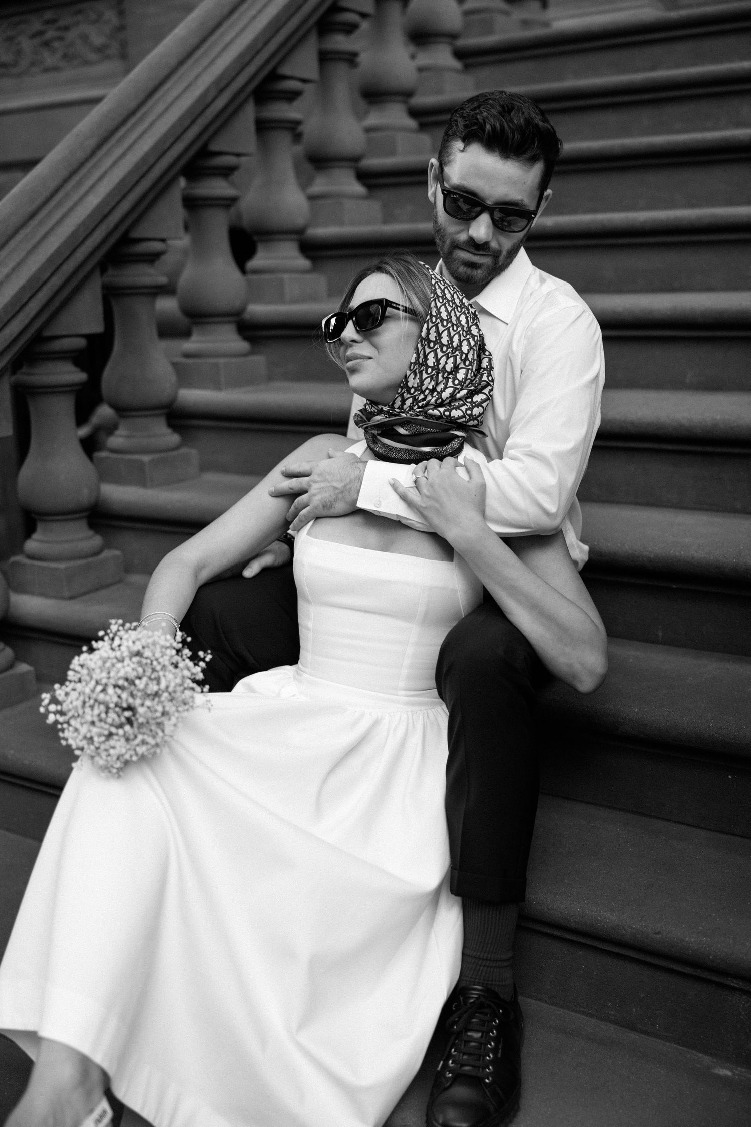 A romantic couple in formal attire sits on stone steps in black and white.