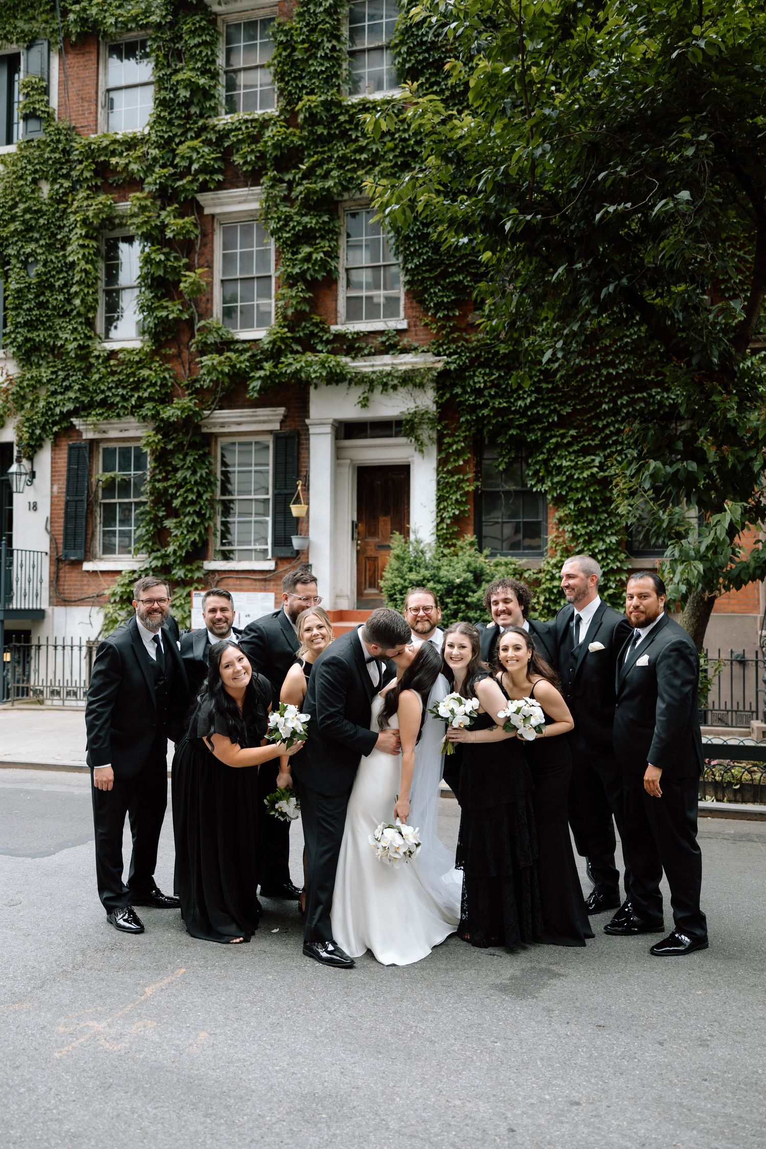 Wedding party in black attire poses together on a tree-lined street.