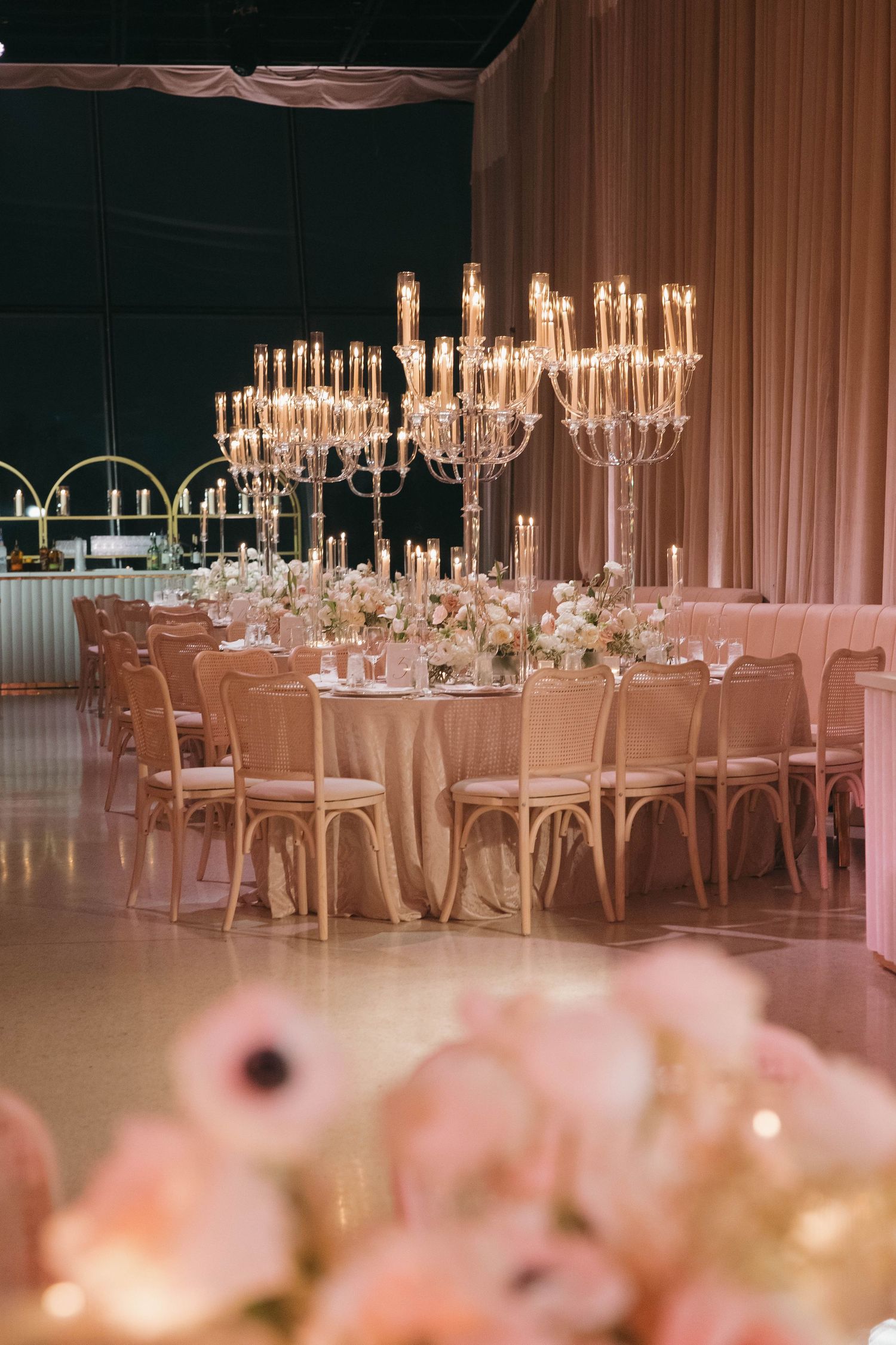 Crystal chandeliers illuminate round reception tables with pink chairs and floral centerpieces.