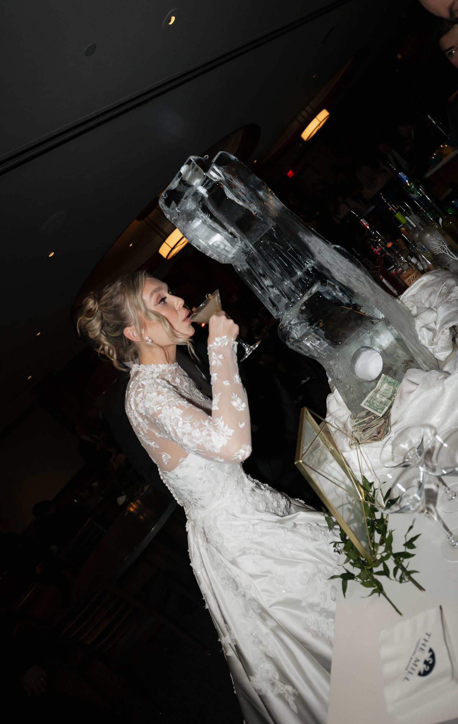 Someone in a white lace wedding gown seated at a reception table with glassware.