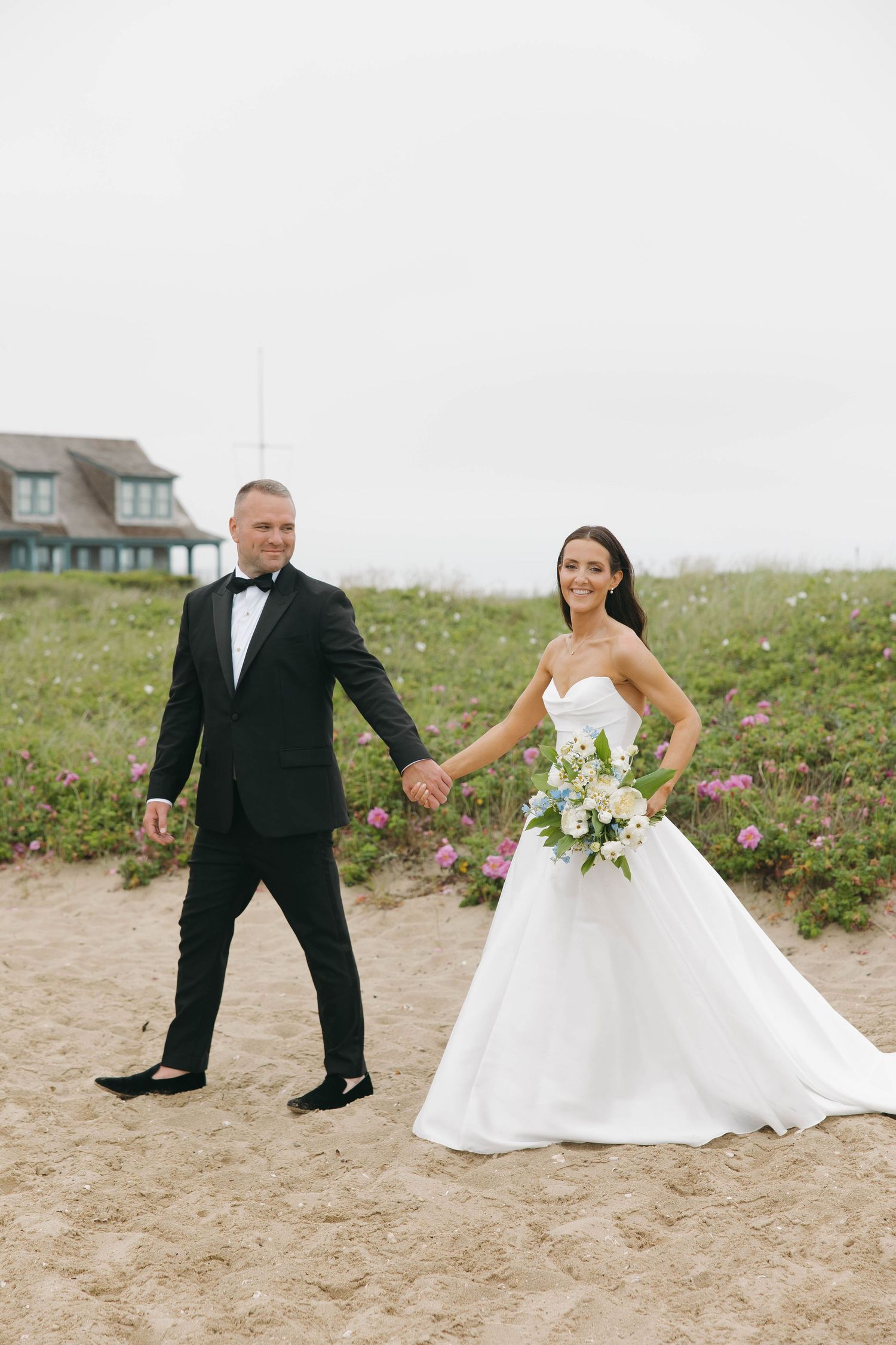 Newlyweds share playful moment on beach with lighthouse in background.