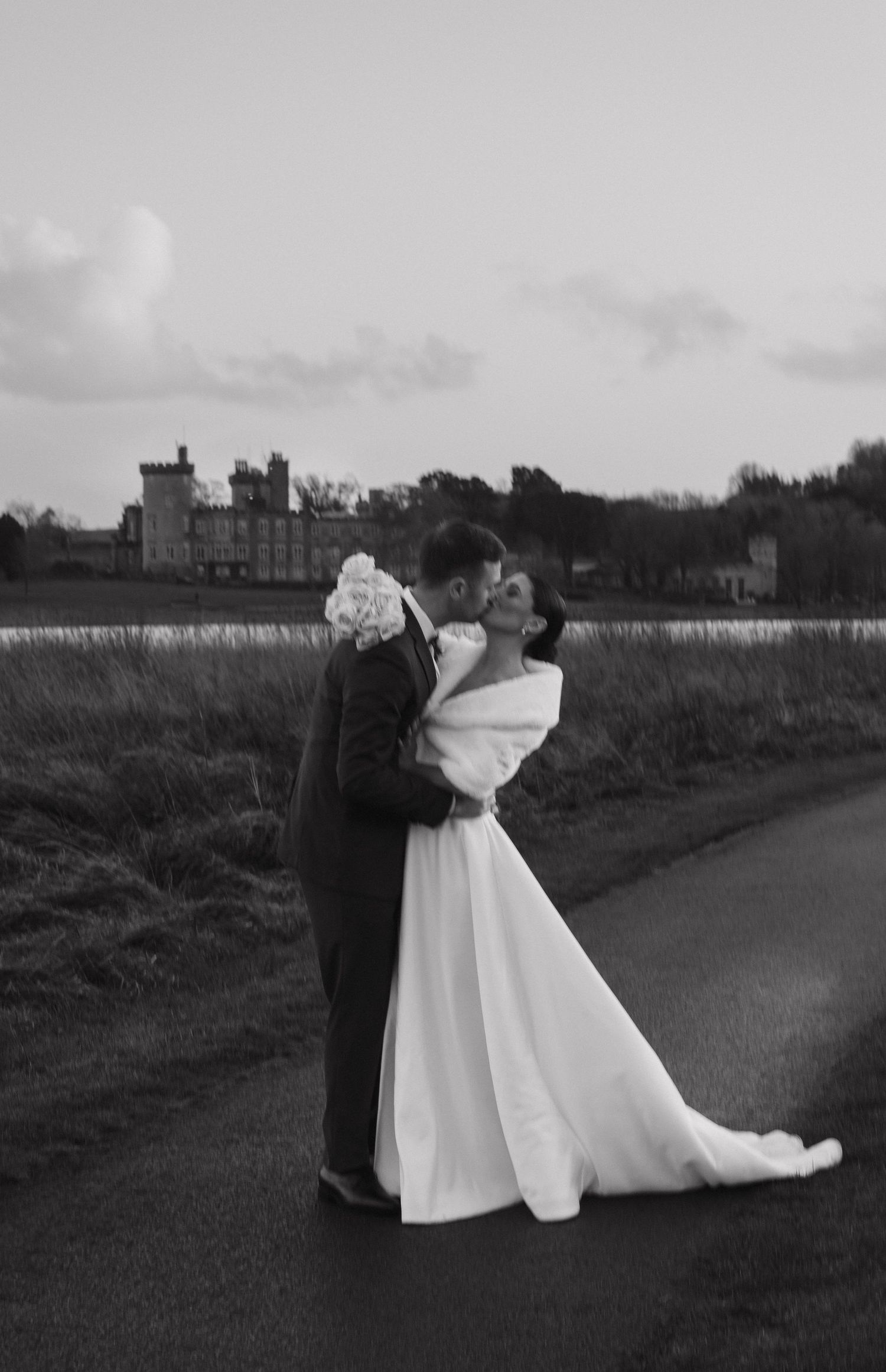 Black and white wedding photo of a couple posing by a castle with water in the background.