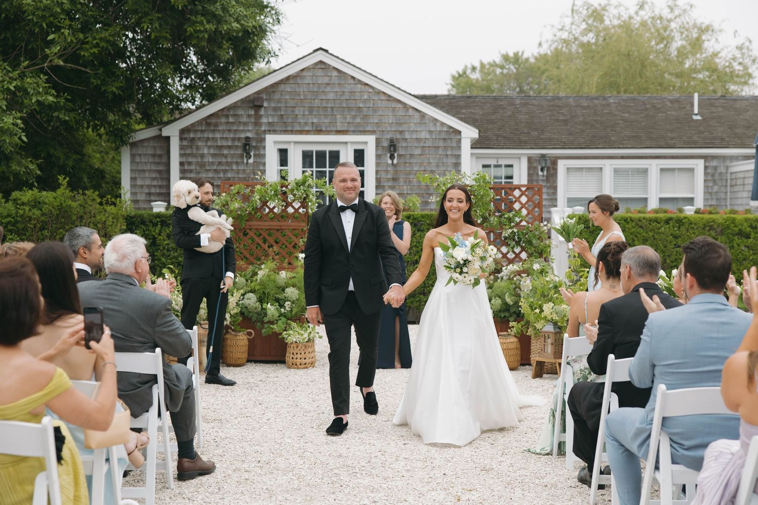 Wide angle view of outdoor wedding ceremony with guests seated on white chairs.
