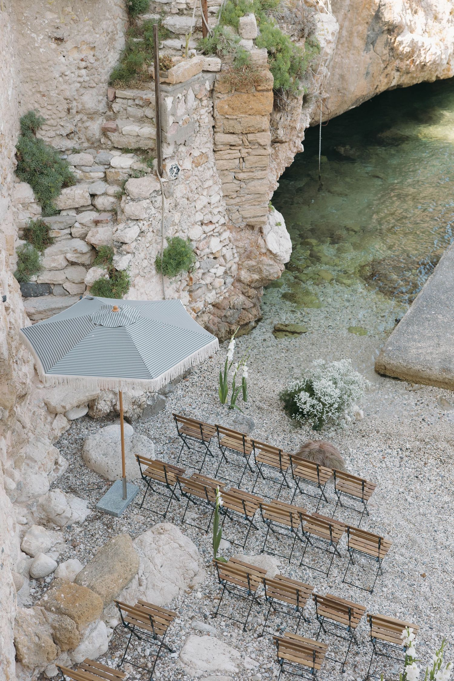 Wedding ceremony setup with wooden chairs and umbrellas nestled within ancient stone ruins by the sea.