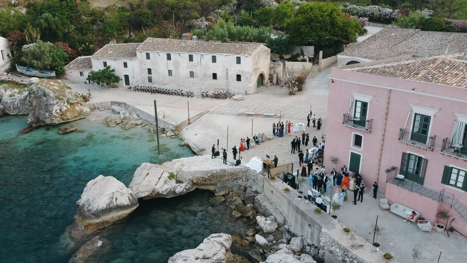 Panoramic view of coastal buildings and gathering crowds along Mediterranean rocky shore.